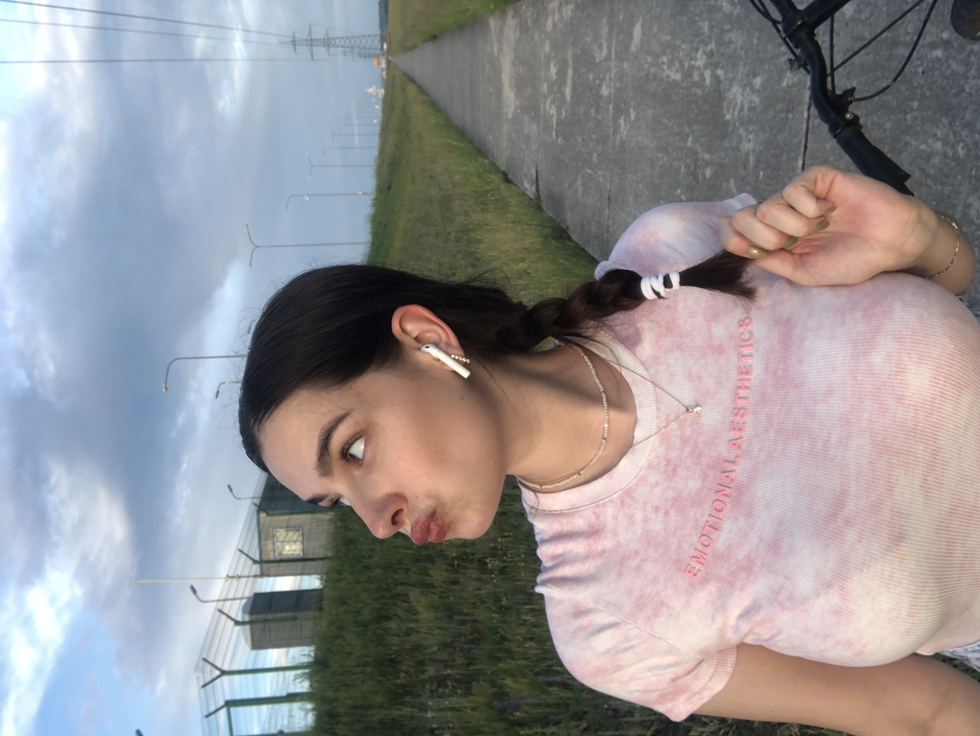 In Noviy, Russia, a young woman with dark hair in a braid, wearing a pink tie-dye t-shirt with the text "EMOTIONAL AESTHETICS," stands on a paved path beside a grassy field. She is looking up and to the left, with her lips puckered. She wears white earbuds and two delicate necklaces. A bicycle is partially visible in the lower right corner. The background features a cloudy sky, a fence, industrial buildings, and utility poles.