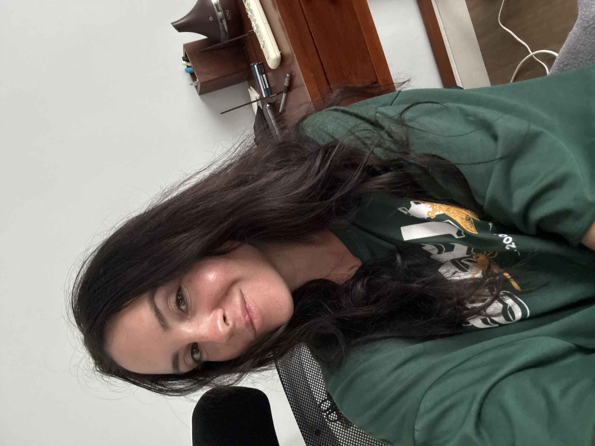 A young woman with long, dark hair is captured in a selfie from the chest up. She is wearing a dark green sweatshirt with a graphic design on the front that includes white and yellow elements, and the number "200". She is seated in what appears to be an office or home office setting, with a portion of a black mesh office chair visible to her left. Behind her, a wooden desk is visible, on which rest a wooden organizer with pens, a dark brown aroma diffuser, a remote control, and what looks like a pen. The background consists of a plain white wall. The lighting suggests an indoor environment.