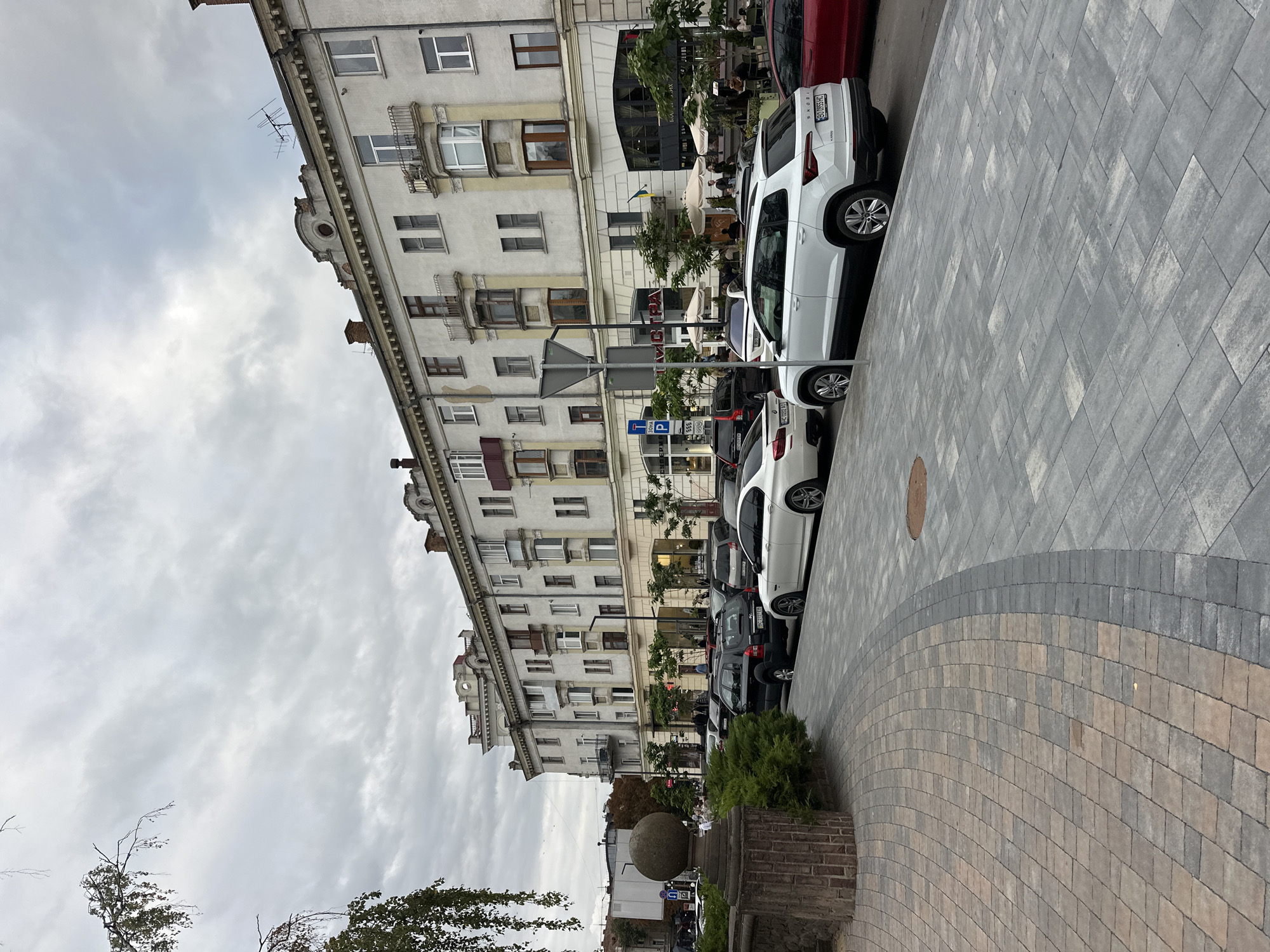 This is a street-level, eye-level, horizontal, full shot of a city street scene in Ternopil, Ukraine, with a focus on the architecture and parked cars. The image captures a row of multi-story buildings with classic European architectural details, such as balconies and decorative cornices, on the right side of the frame. The buildings appear to be a mix of residential and commercial spaces, with storefronts and umbrellas suggesting outdoor seating for a cafe or restaurant on the ground floor. The street in front of the buildings is lined with parked cars, including a white Skoda SUV in the foreground, a white BMW, a black Hyundai SUV, and other vehicles further down the street. The sidewalk area is paved with modern, large gray tiles, with a curved section of smaller, patterned tiles leading towards the foreground. A decorative stone pillar topped with a spherical finial stands on the left side of the frame, adjacent to a manicured green hedge. The sky is overcast and cloudy, suggesting a cool or temperate day. A few sparse tree branches with leaves are visible in the upper left corner. Several people are visible walking on the sidewalk and sitting at outdoor tables. The image appears to be taken during daylight hours.