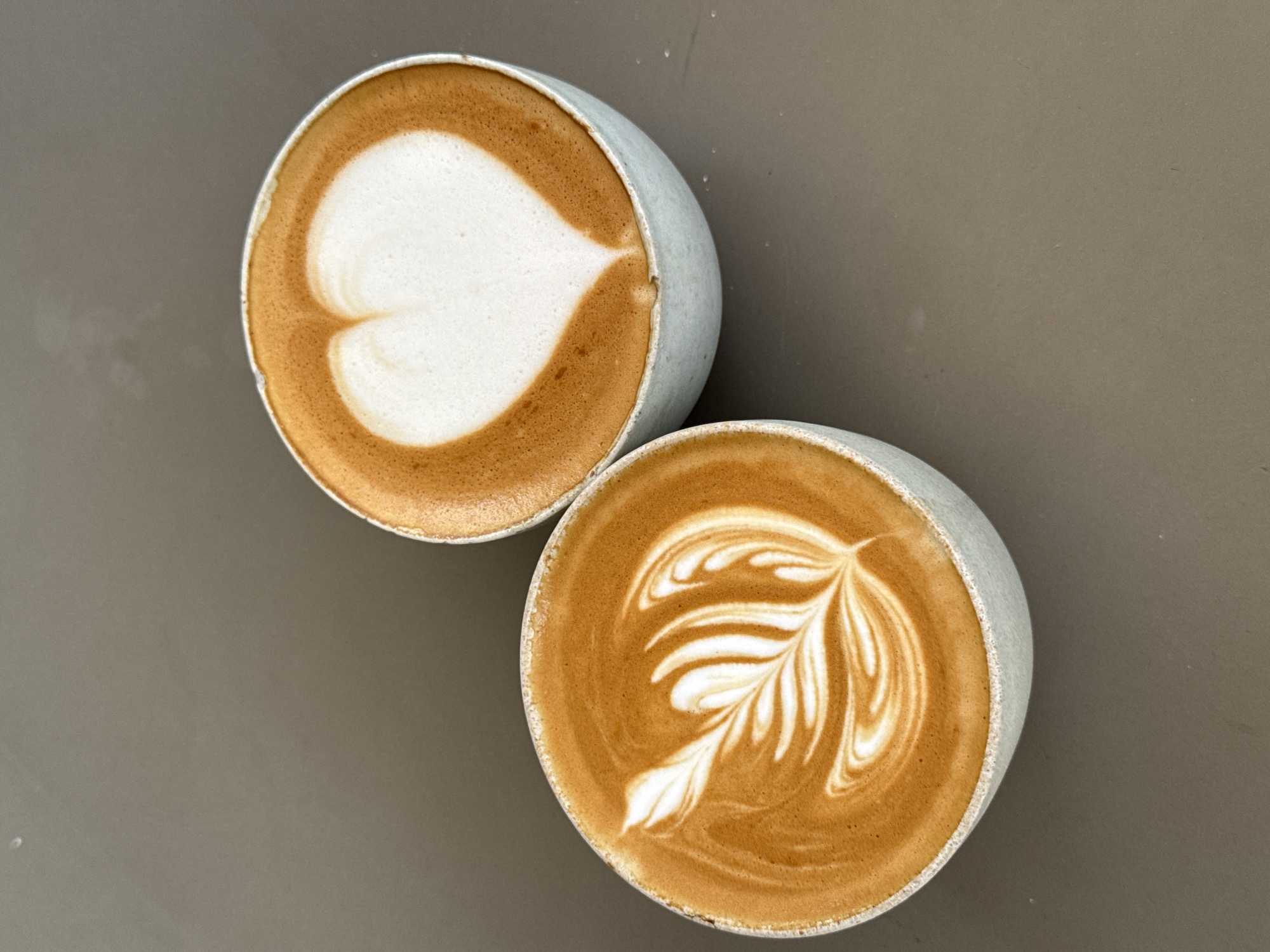 Two cups of latte art coffee are presented on a neutral brown surface. The cup on the left features latte art resembling a feathered leaf pattern in white foam against a rich caramel-colored coffee base. The cup on the right displays a heart-shaped design in white foam, also against a caramel-colored coffee. The cups themselves are light-colored, appearing to be ceramic or stoneware, with a slightly textured exterior. The lighting suggests an overhead source, casting soft shadows. The image is a close-up, overhead shot focusing on the coffee and latte art. The scene is devoid of any human presence or identifiable landmarks to place it specifically in Ko Samui District, Thailand.