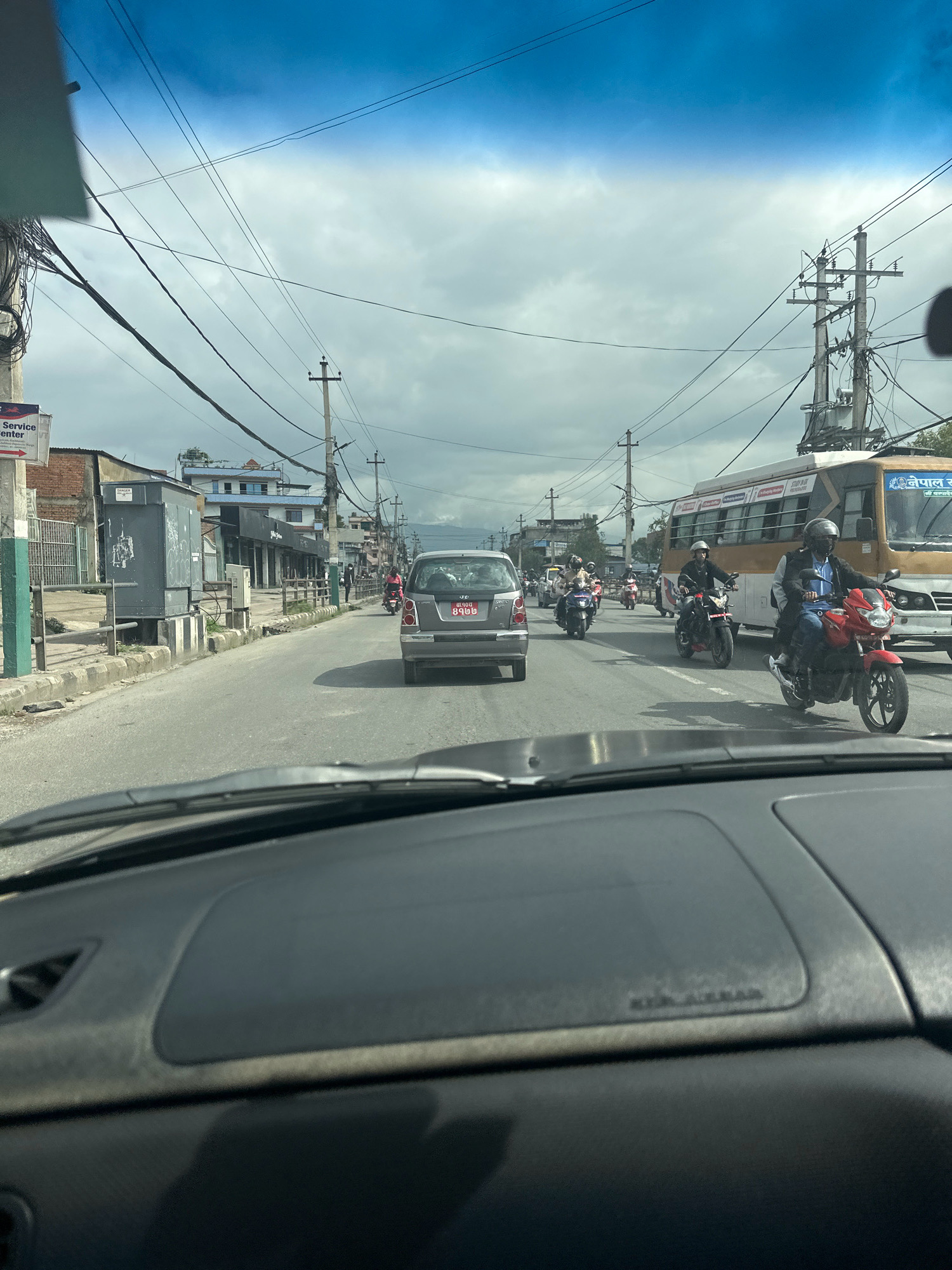 The image depicts a street scene in Kathmandu Metropolitan City, Nepal, captured from the interior of a vehicle. The perspective looks forward, showing the road ahead and the surrounding urban environment. The sky is overcast with scattered clouds.
In the foreground, the dashboard of the car is visible, including the steering wheel area and the passenger side of the dashboard with an airbag cover. The road surface is asphalt, and it appears to be a multi-lane road.
The street is moderately busy with traffic. Several motorcycles and cars are on the road. In the middle ground, a silver car is moving ahead, and to its right, a yellow and white bus is also visible. A red motorcycle with a rider wearing a blue shirt and helmet is in the right lane. Several other motorcycles are scattered throughout the lanes, with riders also wearing helmets.
On the left side of the road, there are buildings, sidewalks, and utility poles with numerous wires. A signpost with red and white text is visible on a pole. A large gray electrical cabinet or enclosure is also present on the left. On the right side, more buildings and utility poles line the street. The overall impression is a typical urban street in Nepal with active traffic and overhead electrical infrastructure.