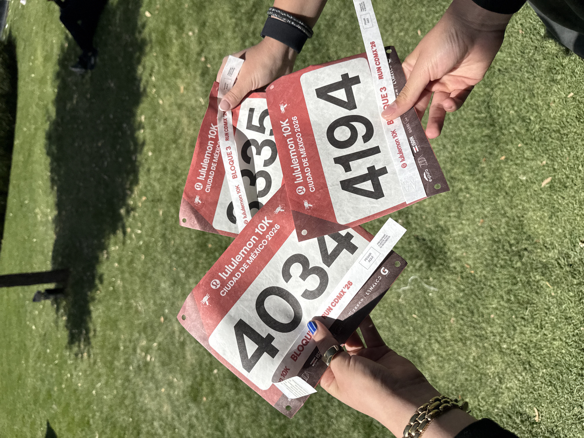 The image shows four running bibs for the Lululemon 10K race in Mexico City, Mexico, in 2026. The bib numbers are 3335, 4194, and 4034. All bibs are Bloque 3. Two people are holding the bibs. One person is wearing a bracelet. The environment appears to be a grassy area.