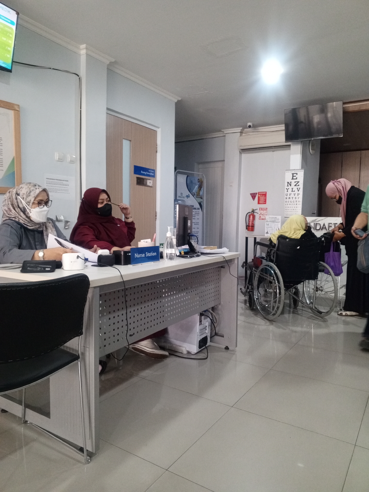 The image depicts a scene inside what appears to be a medical facility in Depok, Indonesia. In the foreground, a nurse's station is visible with two women behind a desk. One woman, wearing glasses and a headscarf, is seated, appearing to be working with documents. The other woman, also wearing a headscarf, is seated and looking to the side. A sign on the desk reads "Nurse Station".
To the right, a person in a wheelchair, wearing a yellow headscarf, is being assisted by another woman in a dark outfit and a pink headscarf. The assistant appears to be adjusting something on the wheelchair or helping the person in it.
In the background, there is a wall with an eye chart displayed vertically. A fire extinguisher is mounted on the wall, and a security camera is also visible. Other elements in the environment include a television screen mounted on the wall, a computer tower under the desk, and a door. The floor is tiled and appears to be reflective. The overall impression is that of an active reception or consultation area within a healthcare setting.