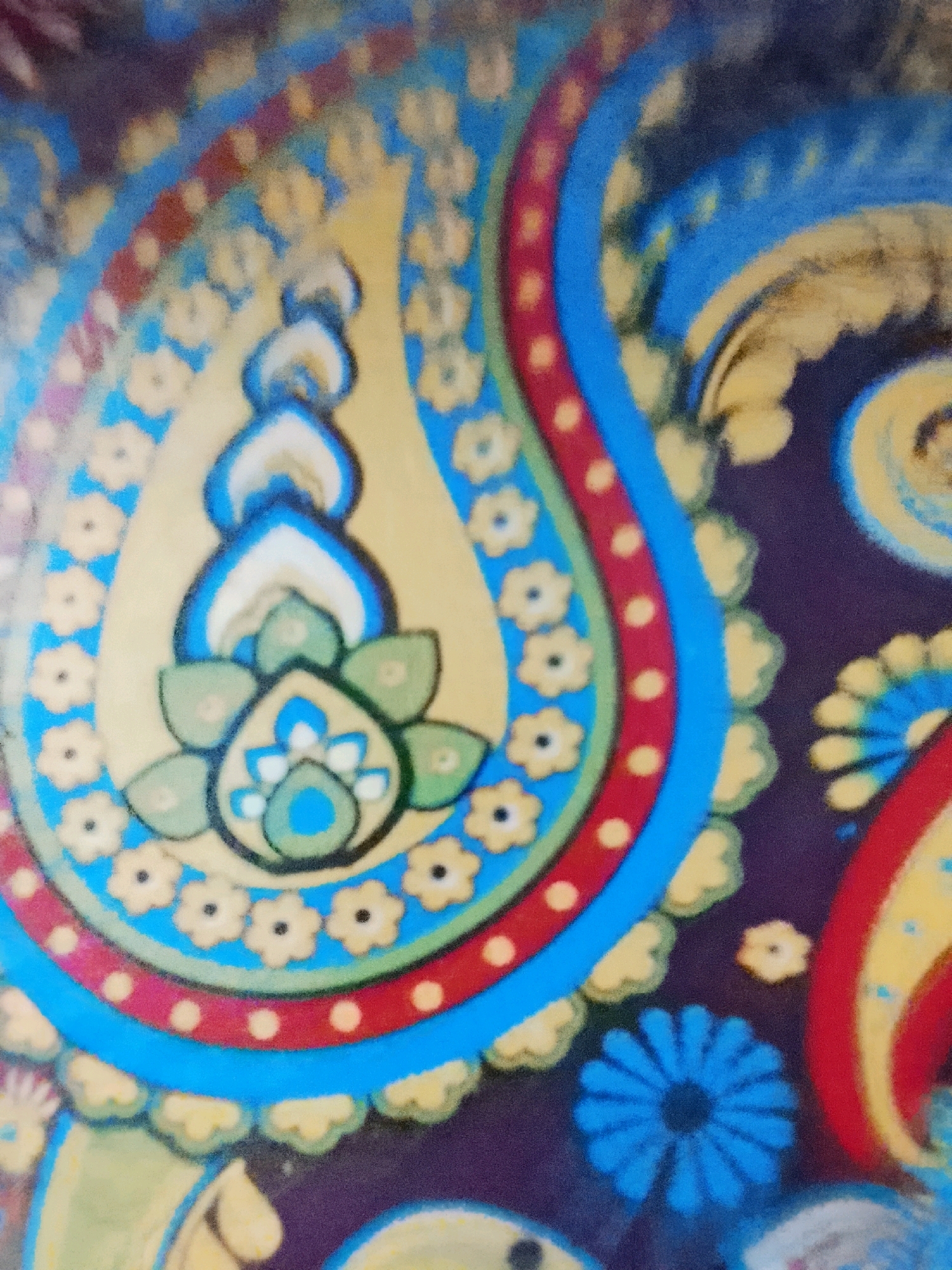 The image displays a close-up of a colorful, decorative pattern, likely a textile or artwork. The dominant design is a stylized paisley motif, rendered in vibrant blues, yellows, reds, and greens against a dark purple background. Small, yellow, flower-like shapes with black centers are scattered around the larger paisley elements. The paisley itself features intricate layering with outlines in black and contrasting colors. There are no people, objects, or discernible environmental details that suggest a specific location in Kuta, Indonesia. The image is purely a visual representation of a decorative design.
