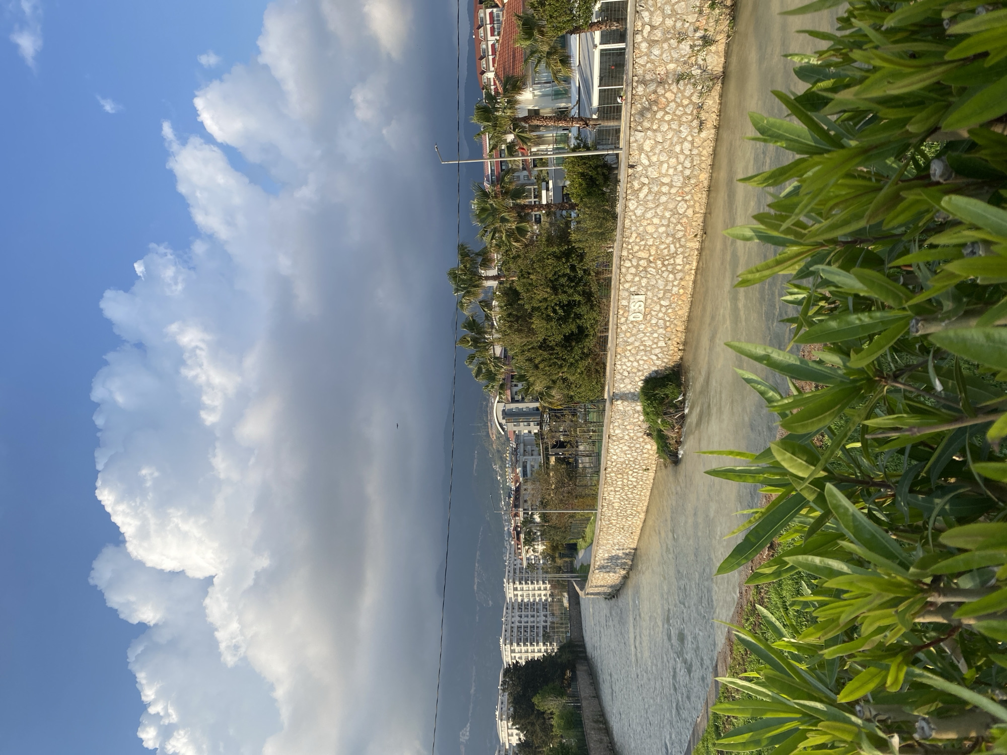 The image presents a view in Alanya, Turkey. A drainage canal or river runs vertically through the frame, bordered by a stone wall. On the right side of the canal is greenery, including foliage. Behind the stone wall, buildings and palm trees suggest an urban landscape. A mountain range is visible in the background. The sky is divided between clear blue with cumulus clouds on the left and ominous dark clouds on the right. The buildings appear to be residential or hotels. There are no visible people in the frame.