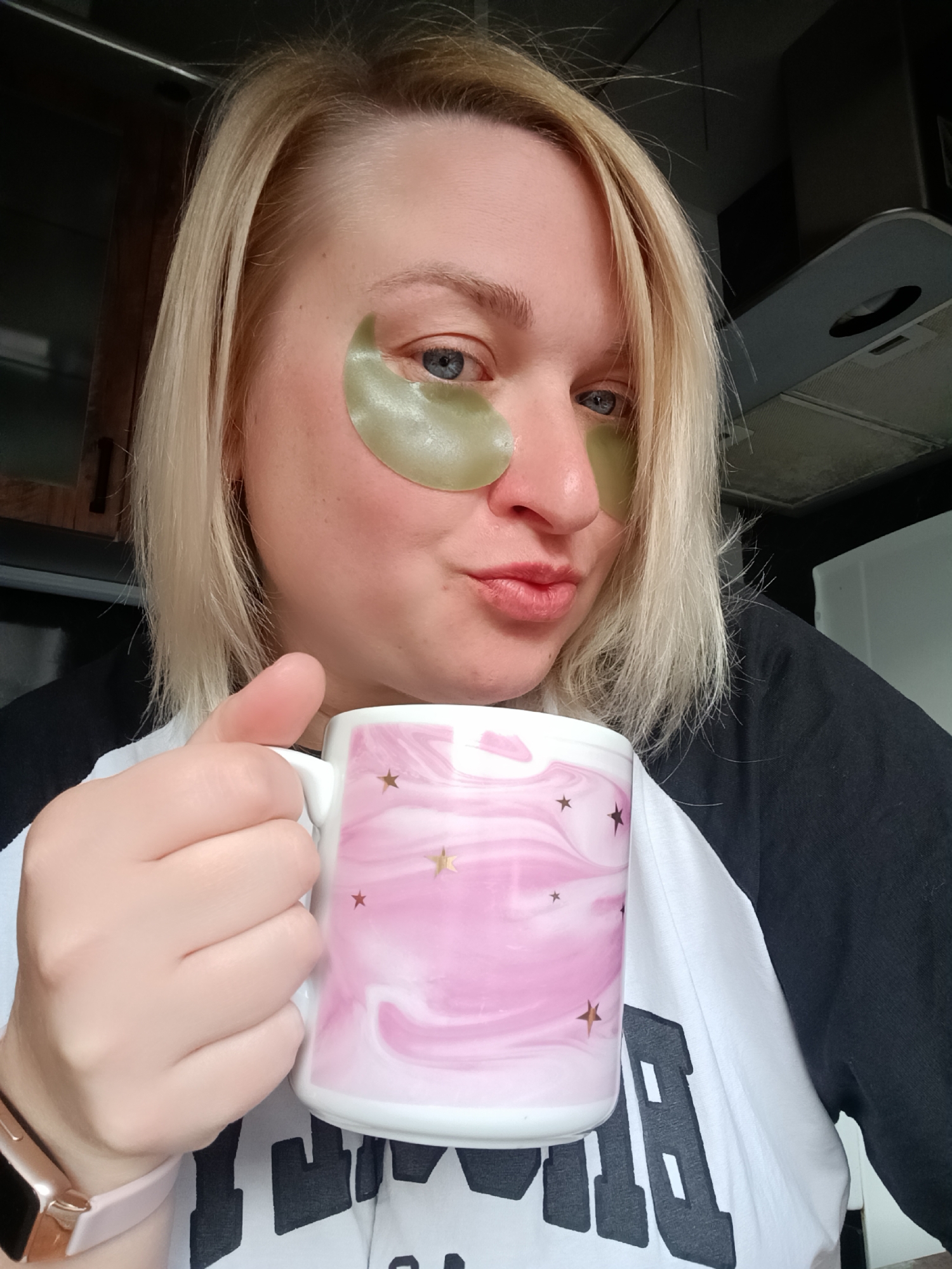 A woman with blond shoulder-length hair is holding a pink and white mug decorated with gold stars in her right hand. She is wearing green under-eye patches and has a slight puckered expression on her lips. She is wearing a black and white t-shirt and a watch with a light pink band. In the background are kitchen cabinets and an overhead range hood. This image was taken in the city of Chaikovsky, Russia.
