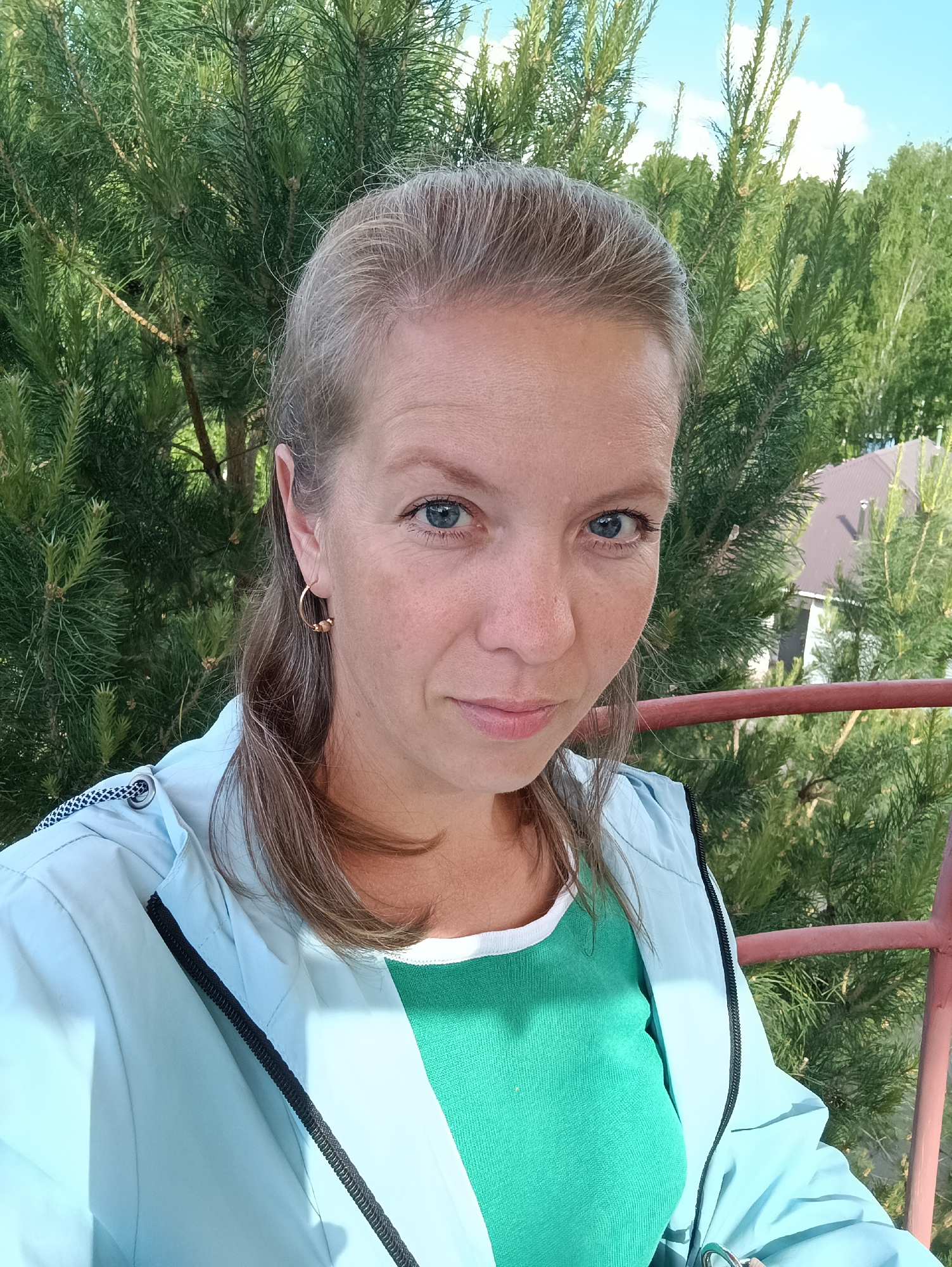 A medium shot captures a light-skinned woman with light hair and blue eyes, facing the camera. She is wearing gold hoop earrings, a green top with a white collar, and a light blue jacket. The background features dense green foliage, possibly pine trees. A red metal railing is partially visible in the foreground. Based on the background foliage, it seems to be summer. The location is Chaikovsky, Russia, based on the available information.