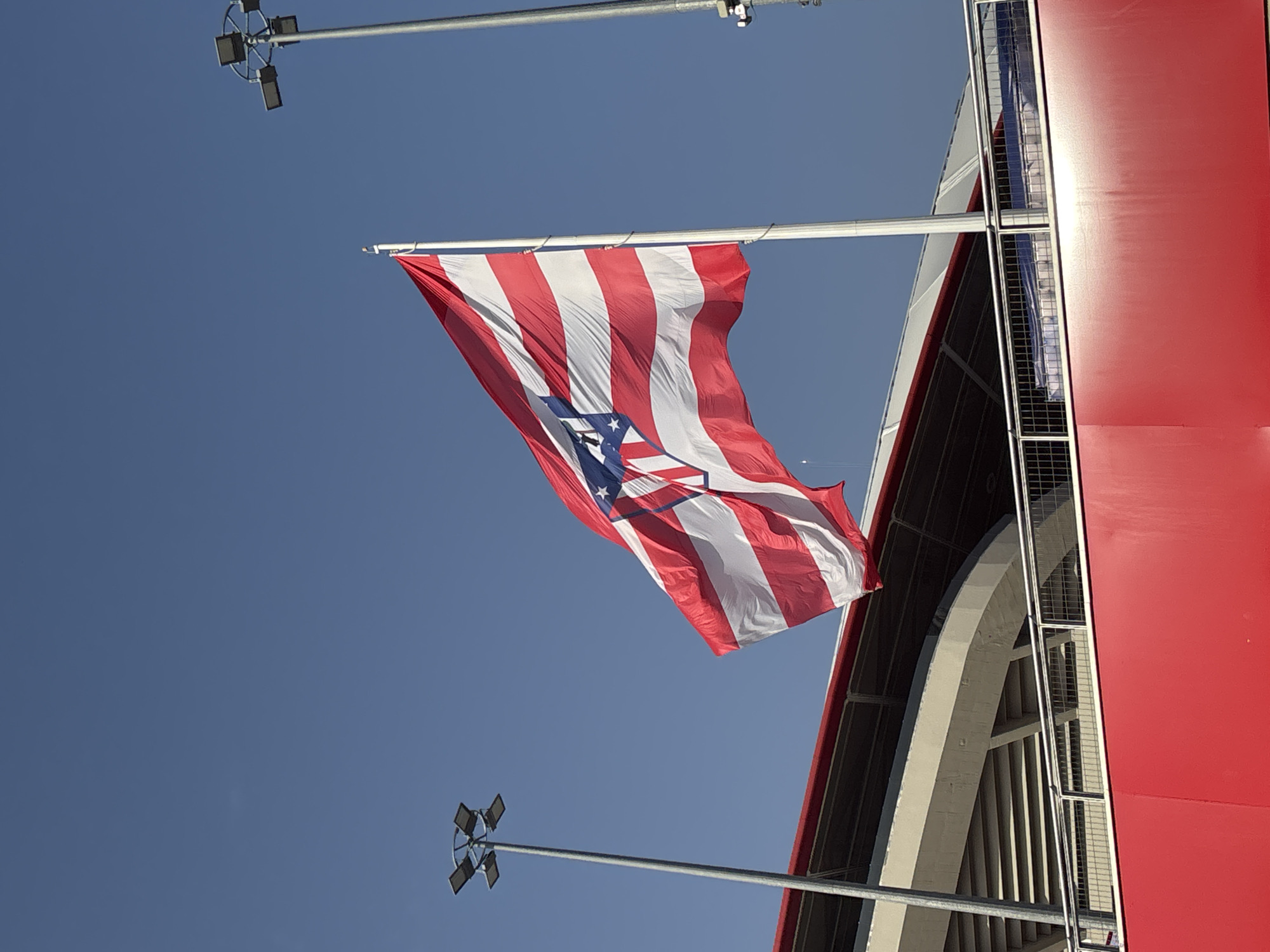 An Atlético Madrid flag is prominently displayed, waving against a clear blue sky. The flag features red and white horizontal stripes, with a blue triangular section on the left side containing a white star. The flag is attached to a tall flagpole. In the background, a portion of a stadium can be seen, including seating areas and a bright red facade. There are also tall lampposts with multiple lights visible, typical of a sports venue. The scene is outdoors, under natural sunlight.
