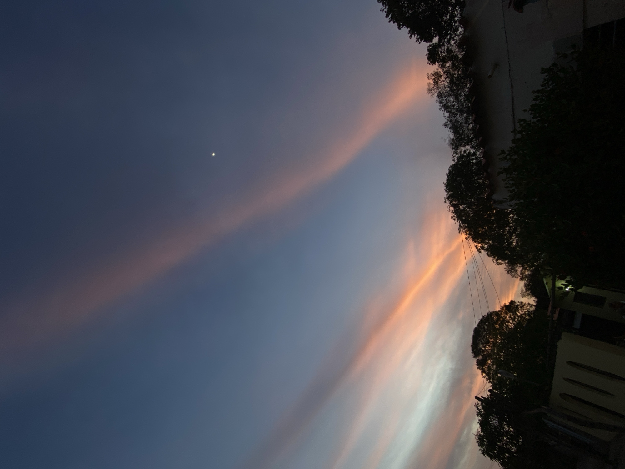 The image captures an evening sky over Juan Griego, Venezuela, at twilight. The sky transitions from a deep indigo-blue at the zenith to lighter blues, purples, and vibrant oranges and pinks near the horizon. Multiple distinct, elongated light rays, consistent with crepuscular rays, traverse the sky diagonally. A small, bright crescent moon is visible in the upper-right quadrant. The lower portion of the frame features dark, silhouetted foliage, primarily trees and dense bushes. Partially visible through and above this foliage are structures and utilities. On the left, a section of a light-colored building or wall with multiple arched openings is discernible. On the right, a portion of another light-colored building with a visible reddish-brown tiled roof is present, with a faint artificial light source emanating from within its structure. Overhead power lines run horizontally across the upper foreground, intersecting with the silhouetted tree line. No individuals are visible.