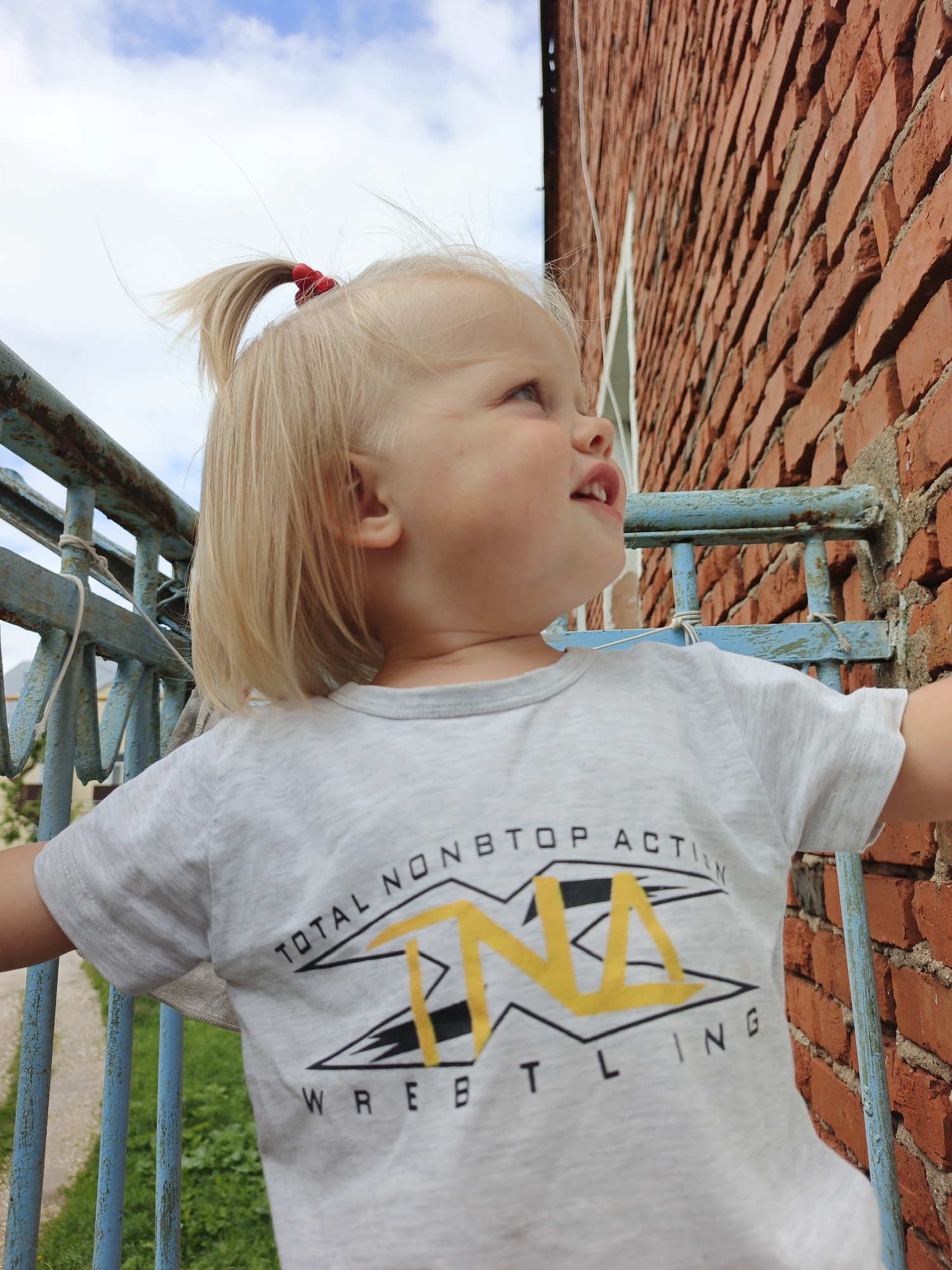 An outdoor medium shot captures a young Caucasian child with blonde hair in a ponytail, wearing a light grey t-shirt with a wrestling logo, leaning against a rusty blue railing. The child is looking upwards and to the right, with their mouth slightly open. Behind the child, a textured red brick wall dominates the right side of the frame, while a cloudy blue sky is visible in the upper left. The blue railing extends from the foreground to the left, with green foliage visible below. The scene appears to be in Medvedyovka, Russia.
