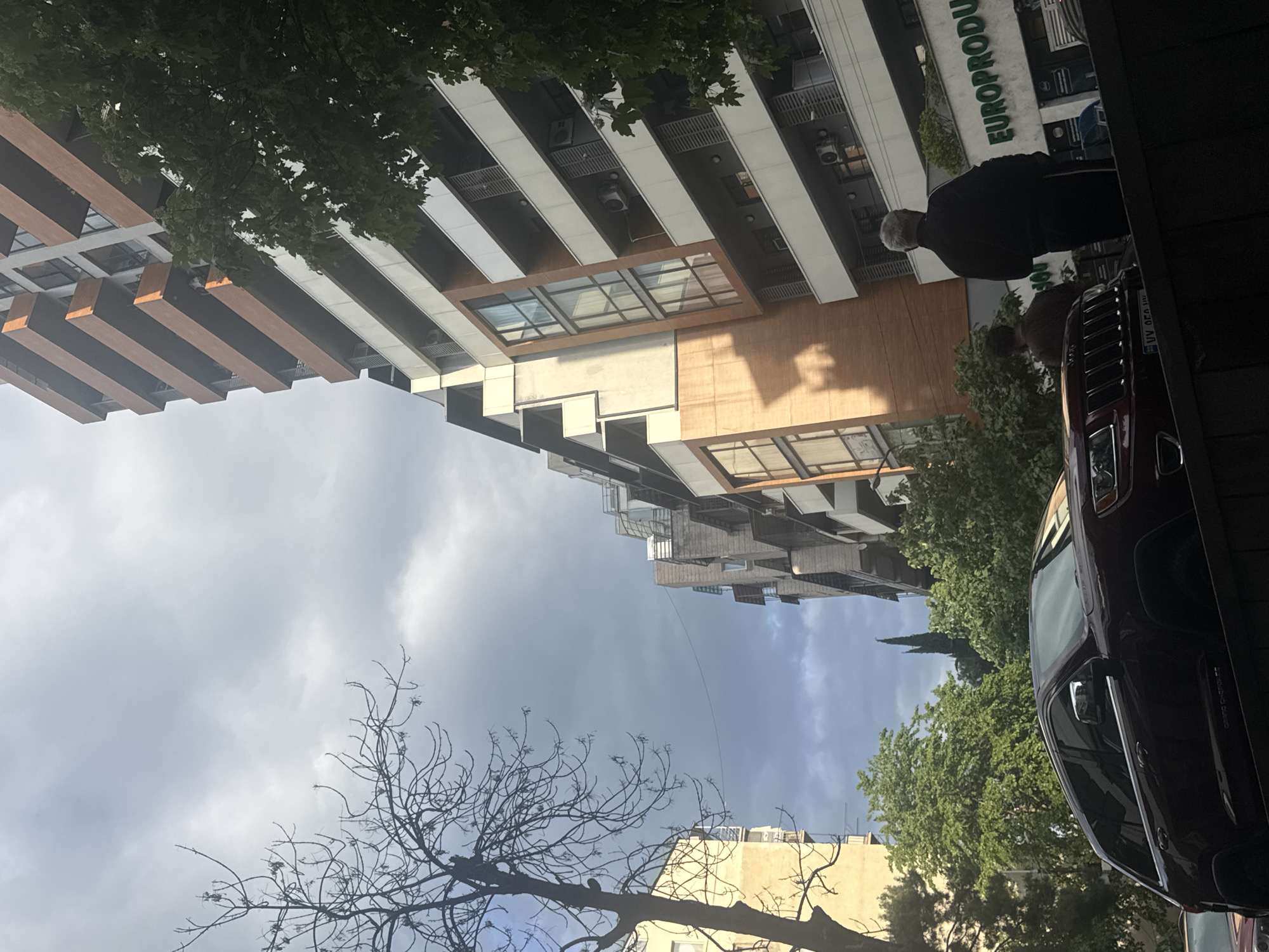 The image shows a street scene in Unknown, Unknown, with a building complex dominating the frame. The buildings are modern, with a combination of white, brown, and glass elements. The lower part of the frame shows a car parked on the right side of the street. The sky is cloudy, with hints of sunlight. There are also trees visible, partially obscuring the buildings. A man is standing near the car, with another person partially visible behind him. The word "EUROPRODU" is visible on the facade of one of the buildings.
