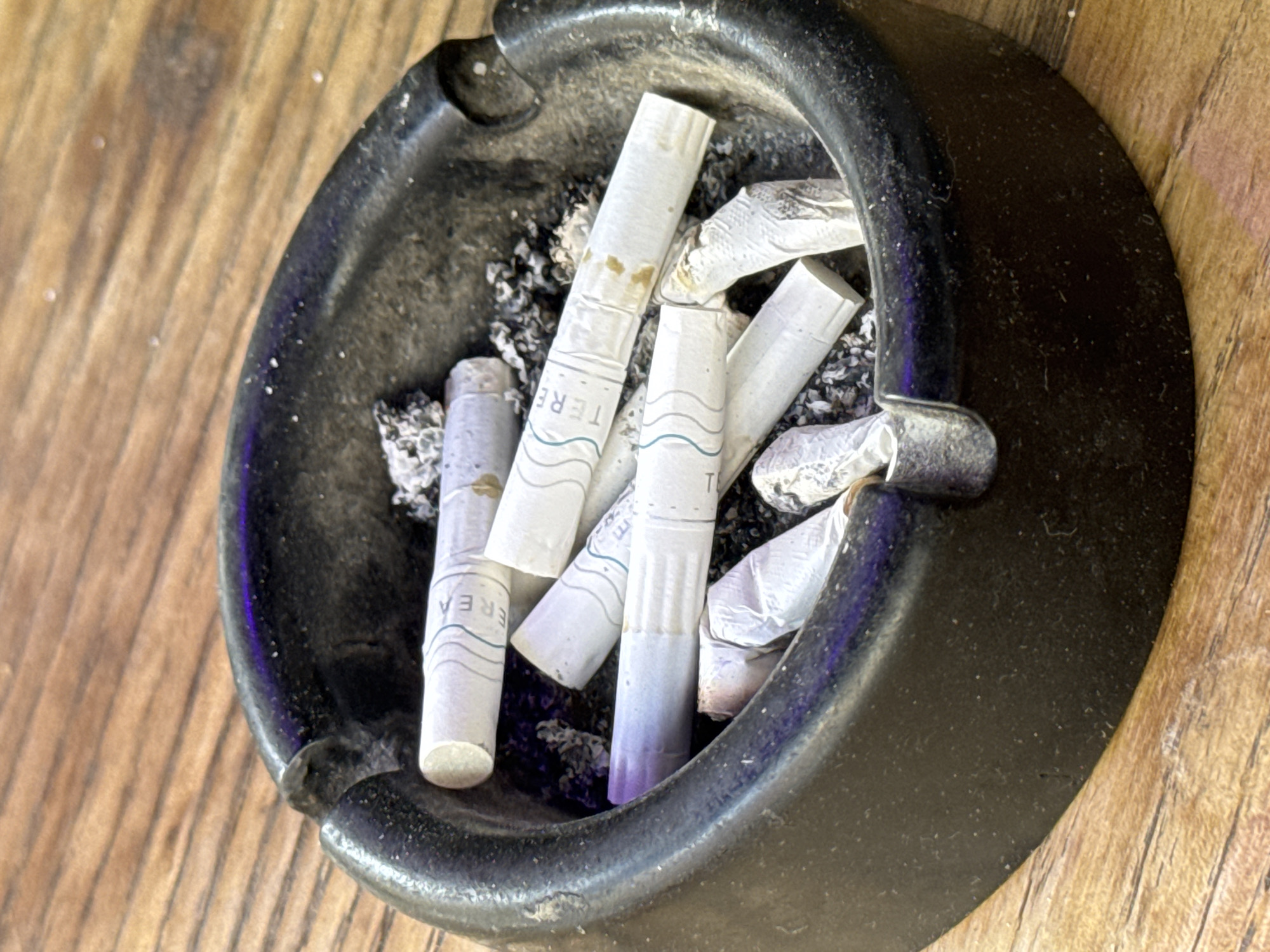 An overhead, close-up shot depicts a dark-colored, circular ashtray filled with numerous used cigarette butts and grey ash, resting on a light brown wooden surface. The ashtray, possibly ceramic or metal, has a textured surface and visible residue of ash and grime on its interior and rim.
Inside the ashtray, several white cigarette-like sticks are prominently featured. Many of these appear to be heated tobacco sticks rather than traditional cigarettes, evidenced by their uniform cylindrical shape and often-intact structure. Visible text on multiple sticks includes parts of the brand name "TEREA" (e.g., "TERE," "REA"), often accompanied by subtle wavy line patterns. Some sticks exhibit yellowish-brown discoloration from tar, while others show hints of blue or purple coloration, suggesting different flavor varieties. Alongside the sticks, crumbled ash and smaller fragments are scattered.
The wooden background features distinct grain patterns and appears to be a table or similar surface, providing a natural, warm contrast to the dark ashtray. The lighting is ambient, highlighting the textures of both the wood and the ashtray's contents. The scene conveys a sense of recent smoking activity, set in Sofia, Bulgaria.