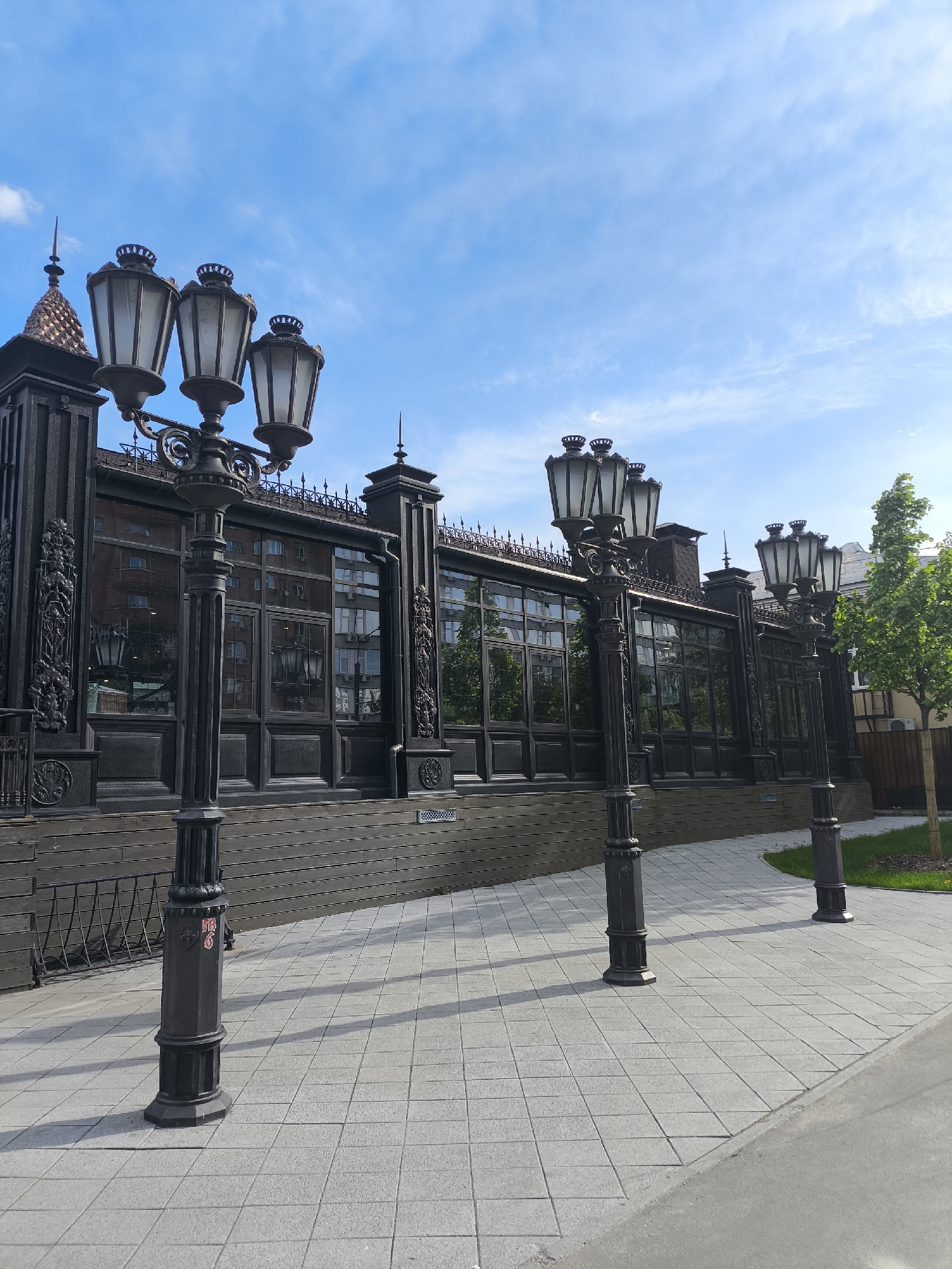 A low-angle, wide shot captures a row of ornate, black street lamps lining a grey-paved walkway in Moscow, Russia. The lamps are mounted on decorative poles, each topped with three glass-enclosed lanterns. Behind the lamps stands a dark-colored building featuring a fence-like wall with repeated window sections. The windows reflect the opposite building, and architectural accents embellish the top of the wall. A strip of greenery runs alongside the walkway, with a small tree planted within. The sky overhead is a pale blue with scattered wispy clouds. The lighting is bright, suggesting a sunny day.