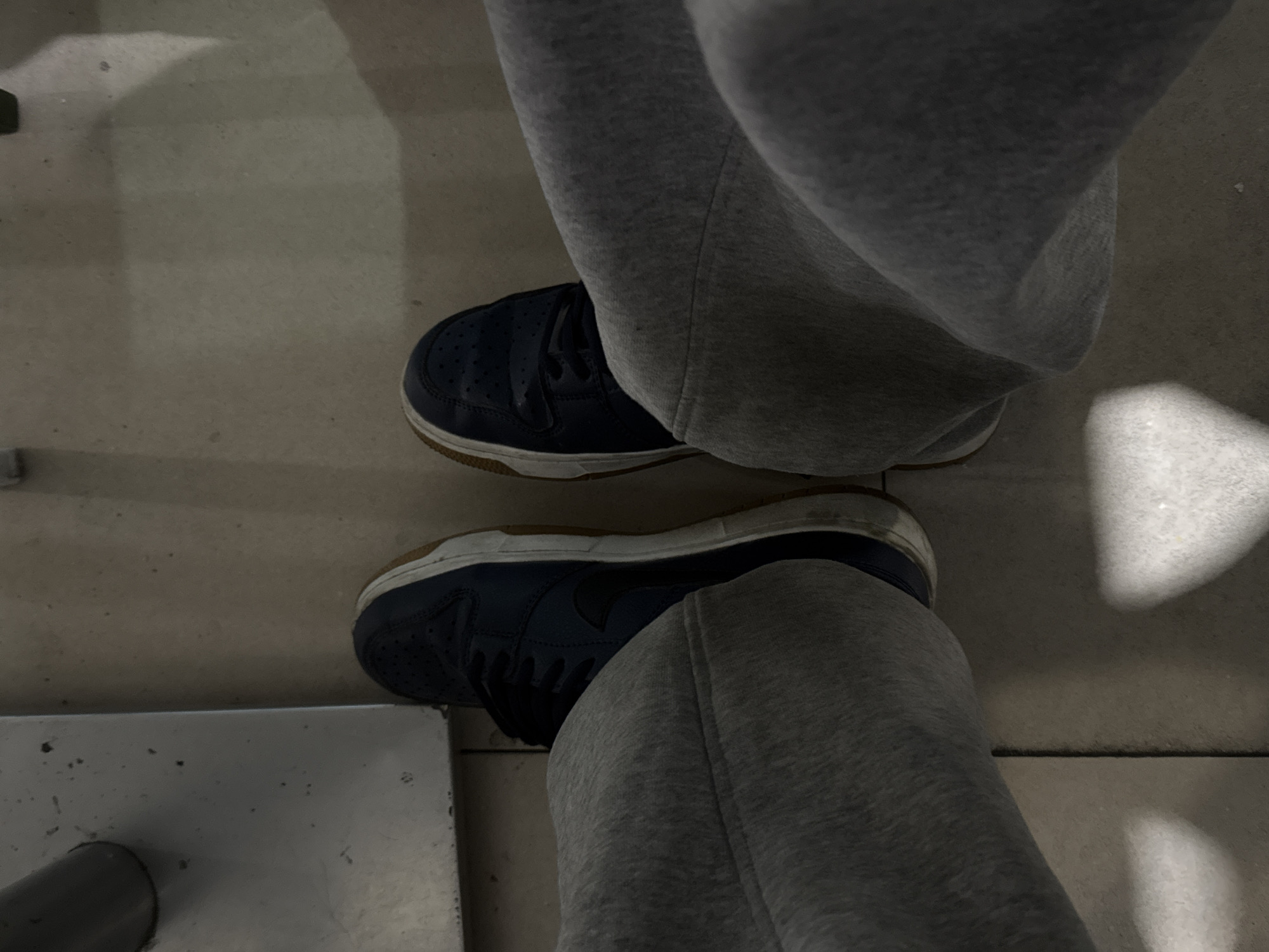 An overhead, slightly angled shot captures the lower legs and feet of a person on a light gray floor, possibly in Yerevan, Armenia. The individual is wearing gray sweatpants or joggers, which are creased and appear relaxed-fit, covering the ankles and upper part of the feet. On their feet are dark blue sneakers featuring white midsoles and light brown or gum-colored outsoles. Subtle perforations are visible on the toe box of the right shoe, and both shoes show signs of wear, particularly on the white midsoles.
The scene appears to be an indoor or covered setting, possibly a hallway or public area, characterized by the plain, light gray floor, which has some minor scuffs or dirt. In the bottom-left corner, a portion of a light-colored, possibly metallic, rectangular object is visible, with a dark gray cylindrical pipe or support extending upwards from it. The lighting is generally dim and diffused, with a distinct bright, irregular patch of light illuminating the floor in the top-right corner, suggesting an overhead light source or reflection. The person's posture suggests they are either standing or sitting with their feet resting on the ground. No specific activity is taking place beyond this stance. No text is visible in the image.