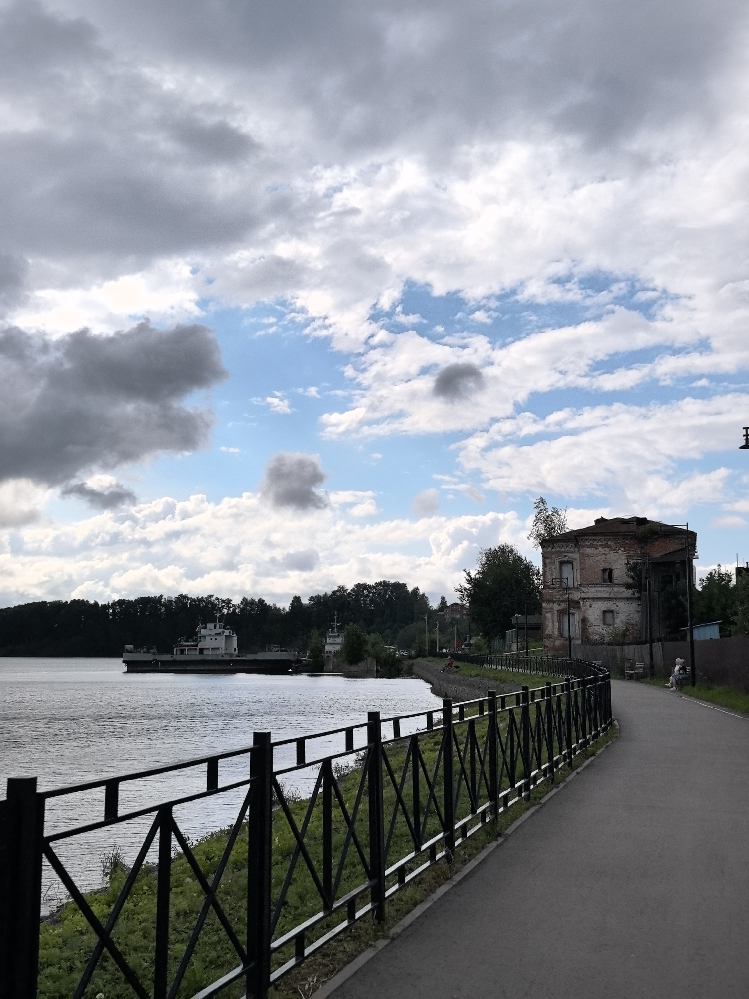 The image depicts a riverside promenade in Dobryanka, Russia. On the left, a wide river occupies the foreground, with a white barge visible in the distance. A black metal fence lines the promenade, separating it from a grassy area that slopes down to the water. The promenade itself is paved and curves to the right, away from the viewer. To the right of the promenade, an old, dilapidated brick building stands, partially overgrown with vegetation. Further along the promenade, two individuals are seated on a bench, observing the scene. The sky is cloudy, with patches of blue visible, and a line of trees forms the background along the riverbank.