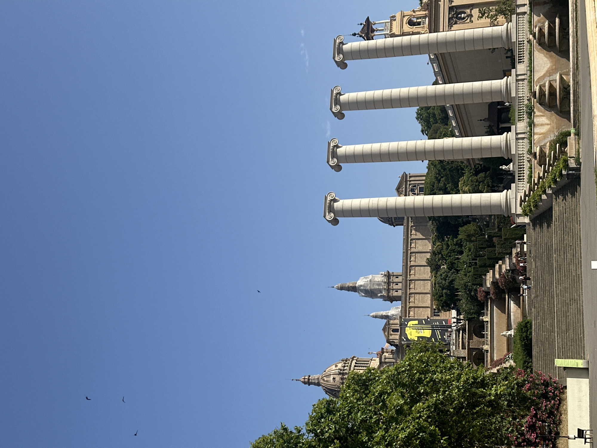 The image shows a bright, sunny day in Barcelona, Spain, likely in the late morning or early afternoon given the clear, blue sky and strong shadows. The scene features a grand architectural ensemble with large, white, fluted columns supporting a building with a classical facade. In the background, more buildings with domes and spires are visible, suggesting a significant historical or cultural site. Lush green trees and flowering bushes add to the vibrant atmosphere, with a tiered garden or plaza area with staircases and fountains visible on the right side. Several people are seen walking on the stairs and near the buildings, appearing to be enjoying the day. The clear sky suggests good weather, and the overall ambiance is one of a picturesque and well-maintained public space. The prominent columns and classical architecture are strong visual cues for the location, possibly indicating a monument or museum. One large yellow banner with black text is visible in the mid-ground, though the text is not clearly legible. The image is taken from a slightly elevated position, looking towards the architectural complex and sky.