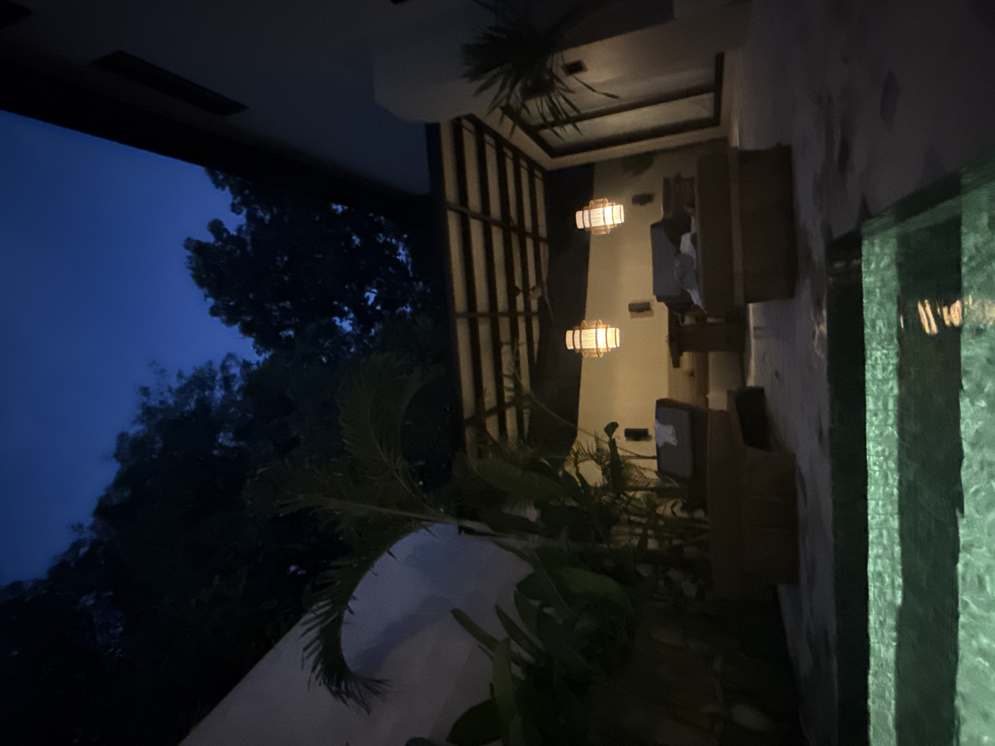 The image shows an outdoor patio area in Kuta Selatan, Indonesia, at night. The area includes a seating arrangement with what appears to be a couch and chairs. There are two cylindrical lanterns hanging from the ceiling, providing light. Next to the patio is what seems to be a swimming pool or reflecting pool, indicated by the green, rippled surface. The surrounding environment includes trees and other vegetation. The sky is dark blue, indicative of dusk or night.