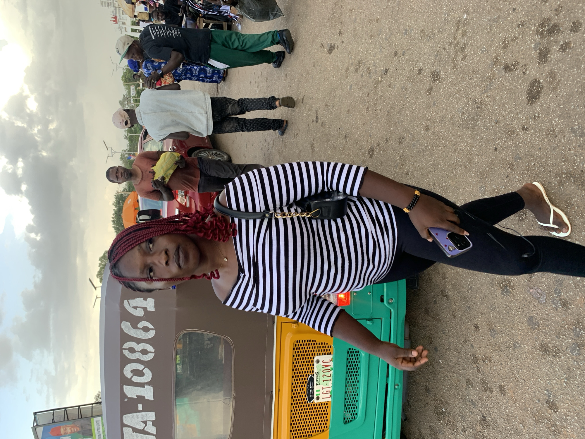 In Jos, Nigeria, a woman with vibrant red braided hair walks on a paved street, her gaze directed forward. She wears a black and white horizontally striped off-the-shoulder top and black leggings, accessorized with a beaded bracelet on her left wrist and a small black purse with a gold chain strap slung over her shoulder. In her right hand, she holds a purple-cased mobile phone.
Behind her, a brightly colored auto-rickshaw, painted in green, yellow, and brown, is parked. The brown section displays white lettering that reads "TA-10864". A Nigerian license plate, "LGT-120VC", is visible on the yellow part of the auto-rickshaw.
In the background, other individuals are present. A man in a rust-colored t-shirt holds a yellow plastic bag. Another man wears a light-colored vest over dark pants, and a third man in a black t-shirt and green pants stands to the right, carrying a large black bag. The scene unfolds under a cloudy sky, with streetlights and billboards visible in the distance, suggesting an urban environment in Jos.