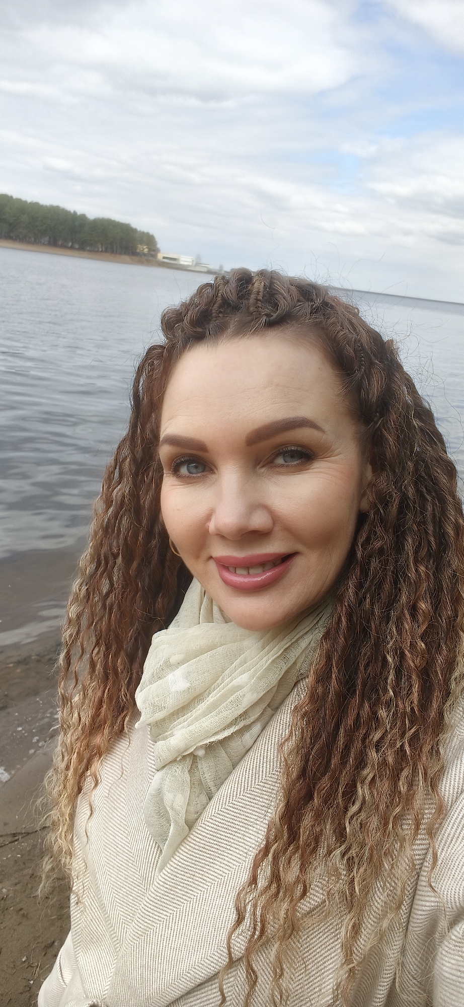 The image is a selfie of a woman with long, curly auburn hair and light skin. Her hair is braided at the crown and she has blue eyes. She wears light makeup, including lipstick. She is wearing a light-colored herringbone coat and a light-colored scarf.
The background shows a body of water, likely a river or lake, with trees visible on the opposite shore. The sky is overcast. Based on the location information, this is likely a scene on the banks of the Kama River within the city of Chaikovsky, Perm Krai, Russia.
