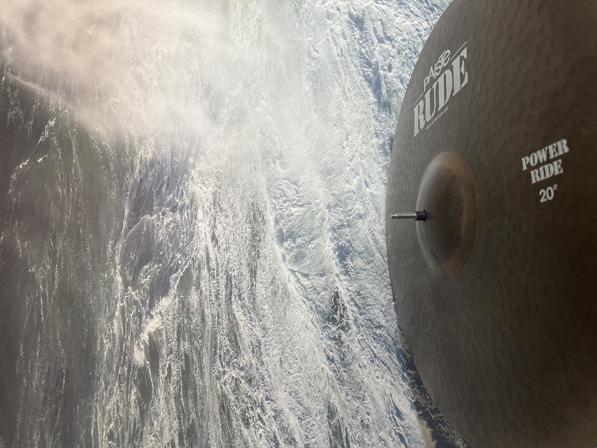 The image shows a close-up view of a dark-colored cymbal, likely a ride cymbal, with the brand name "Paiste" and "RUDE POWER RIDE 20" visible in white lettering. The cymbal is positioned in the foreground, with its dome-shaped central area and a threaded screw at the apex clearly visible.
In the background, a turbulent ocean or large body of water with whitecaps and frothy waves is depicted. A bright, hazy light source, possibly sunlight or spray, illuminates the water on the right side of the frame, creating a strong contrast.
There is no visible evidence or indication of the city of Nevinnomyssk, Russia, in this image. The scene appears to be set at sea or a large coastal area. There are no people or specific actions or interactions being depicted.