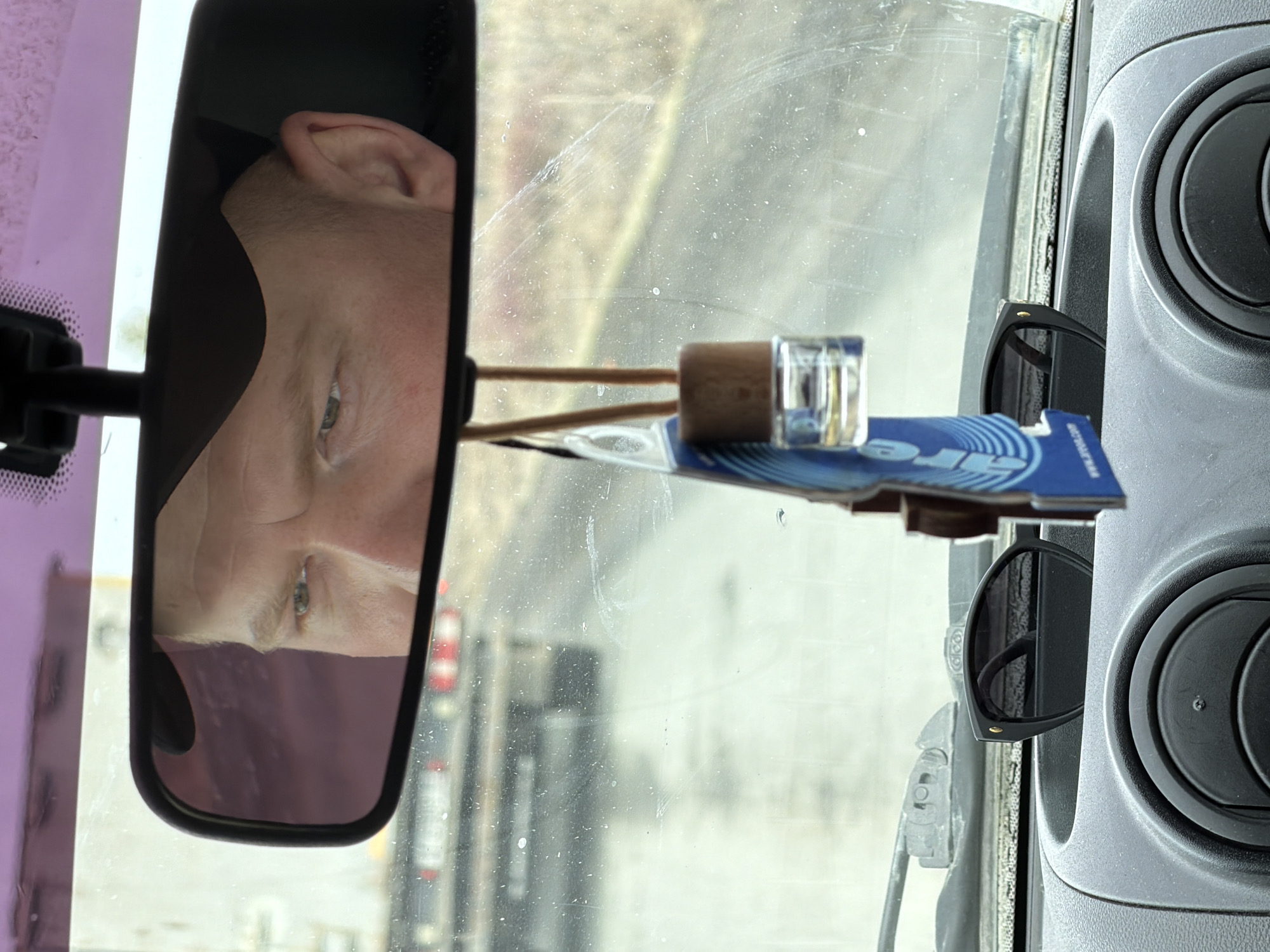 The image shows the interior of a vehicle. Reflected in the rearview mirror is the face of a man. On the dashboard is an air freshener and a pair of sunglasses. The windshield is dirty. Through the windshield, the blurred image of another vehicle can be seen. There is no information to identify the location in Bashur, Russia.