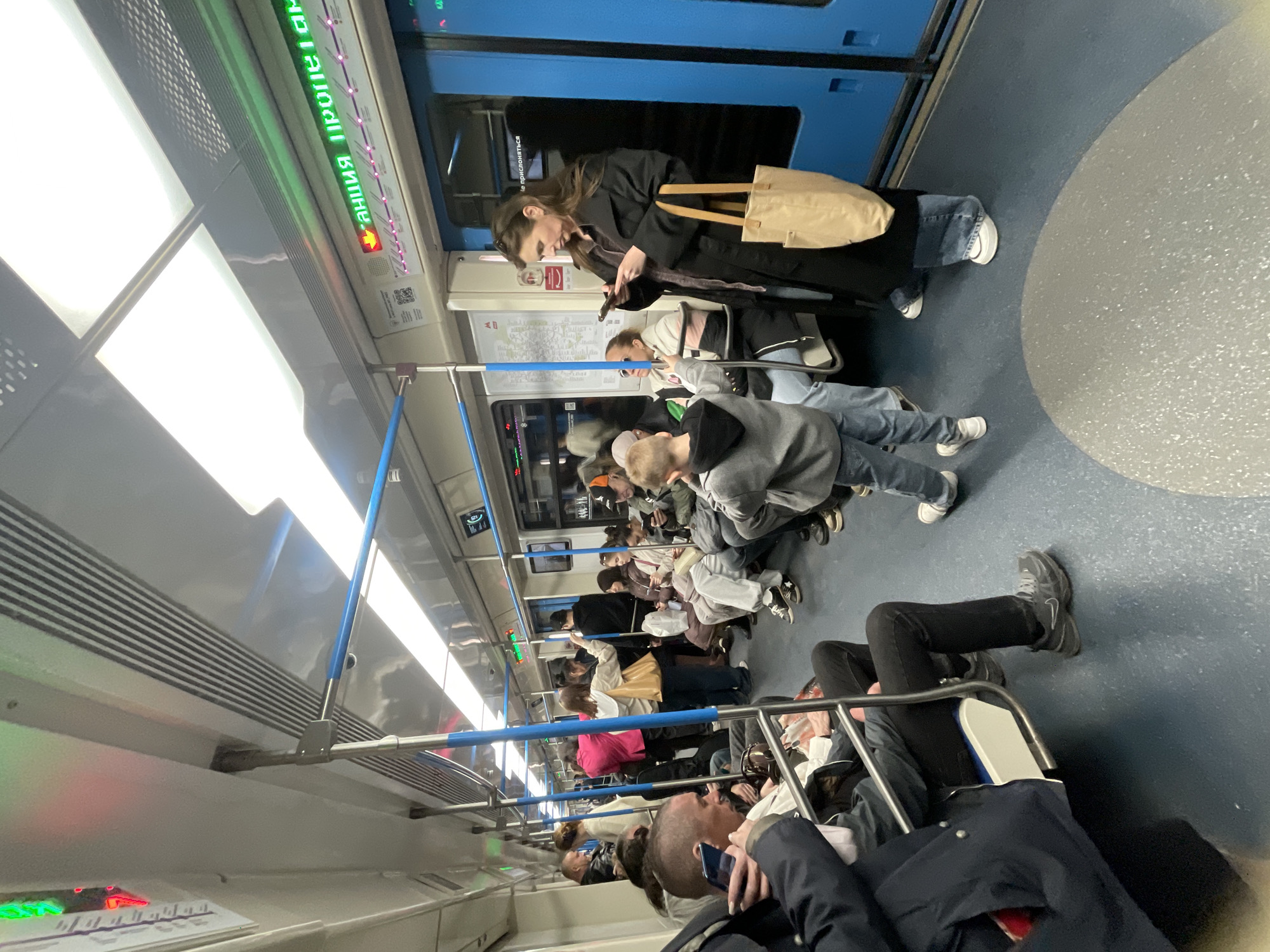 The image depicts the interior of a subway car, likely in Moscow, Russia, given the Cyrillic script on the electronic information board overhead. The car is occupied by numerous people of varying ages and clothing styles. Most are standing or sitting, with several individuals looking at their mobile phones. There are metal handrails, blue in color, running along the length of the subway car. The floor is a blue-gray color with a speckled texture. The walls are a light gray. The doors are a blue tone.