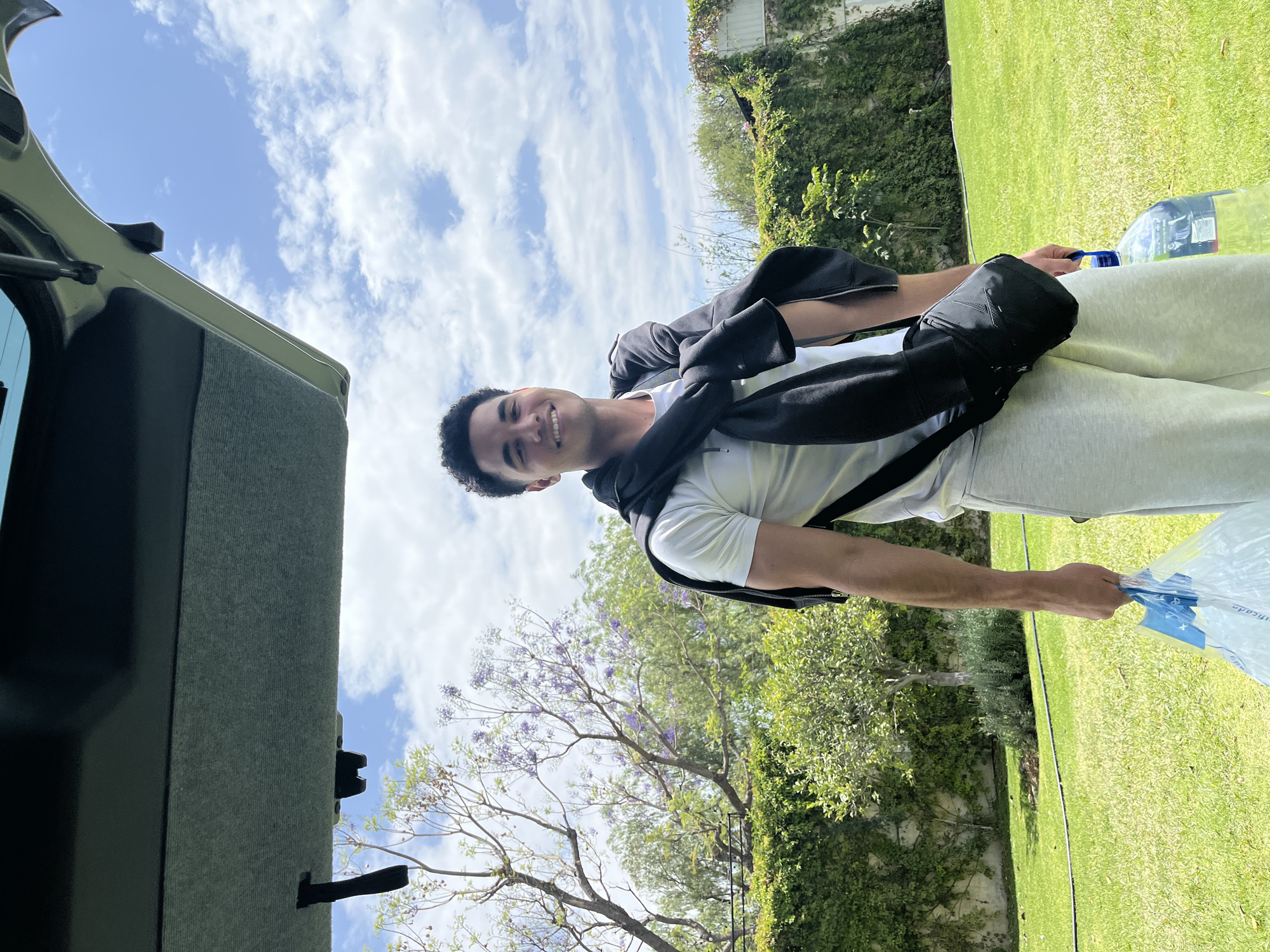 A young man stands in a grassy yard with green bushes, a tree with purple flowers, and a cloudy blue sky. He is wearing a white t-shirt, light-colored sweatpants, and a black jacket is draped over his shoulders. He is smiling and holding a large water bottle. The open trunk of a car is visible on the left side of the image. The location is likely Puebla, Mexico, based on details of the environment.