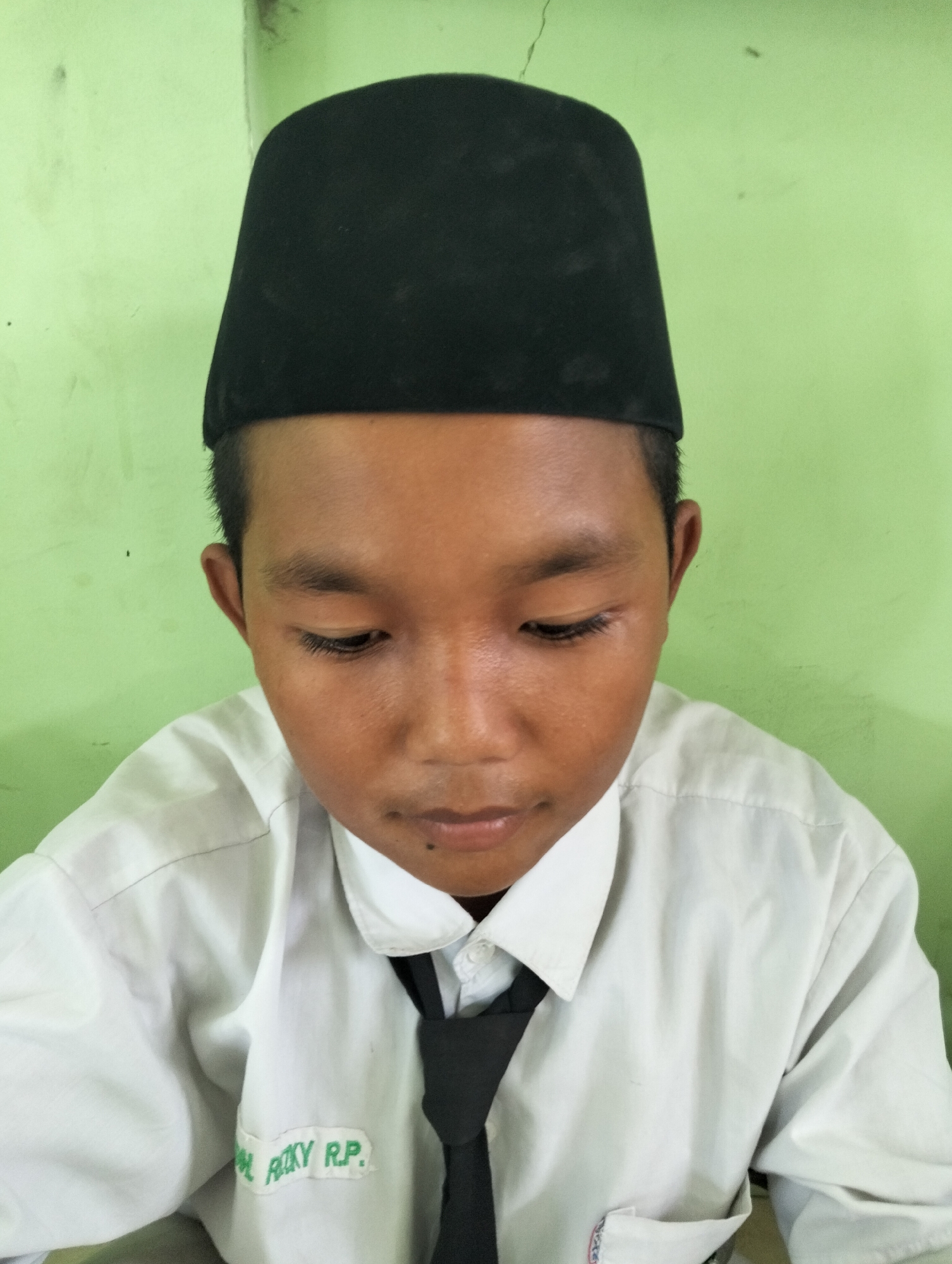 A close-up, head-and-shoulders portrait shows a young boy wearing a black peci (a type of cap) and a white collared shirt with a black tie. The boy is looking down and to his right. On his shirt, there is a rectangular patch with green lettering that reads "ROKY R.P.". The background is a plain, light green wall. The image appears to have been taken indoors, possibly in a classroom or a home in Kranji, Indonesia, given the attire and the general aesthetic.