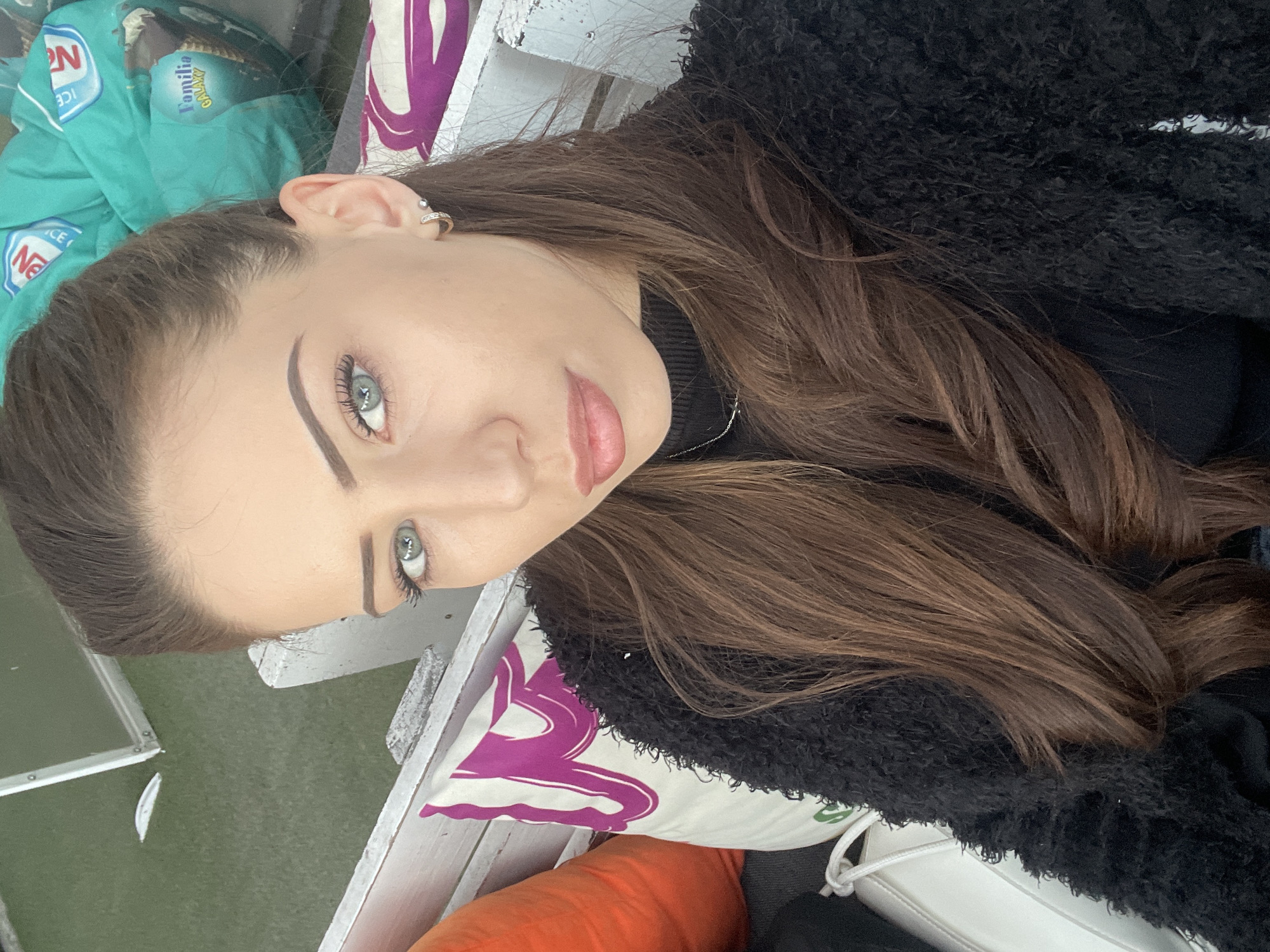 A close-up, slightly rotated portrait captures a young woman seated in an outdoor or semi-outdoor setting, likely in Veliko Tarnovo, Bulgaria. The woman is the primary subject, looking directly at the camera with a neutral, calm expression. She has long, dark brown hair, light blue-grey eyes, and naturally pink lips. She is wearing a textured, fluffy black jacket or coat over a dark top. A small, jeweled hoop earring is visible in her right ear.
The scene suggests a relaxed atmosphere, possibly a cafe or lounge patio. She appears to be sitting on a white wooden bench or chair. To her side, a pillow with a striking purple and white abstract pattern provides a pop of color. Below this, an orange fabric item, possibly another cushion or blanket, is visible. In the background, behind her head, a turquoise-green fabric item is partially visible. This item clearly displays the text "NESTLÉ ICE" and "FAMILIA GALAXY," suggesting it is a branded cooler bag, blanket, or similar accessory. Further back, a patch of green ground or artificial turf can be seen. The lighting is soft and diffused, indicative of an overcast day or indirect light. A white shoe with laces is also partially visible in the bottom right corner.
