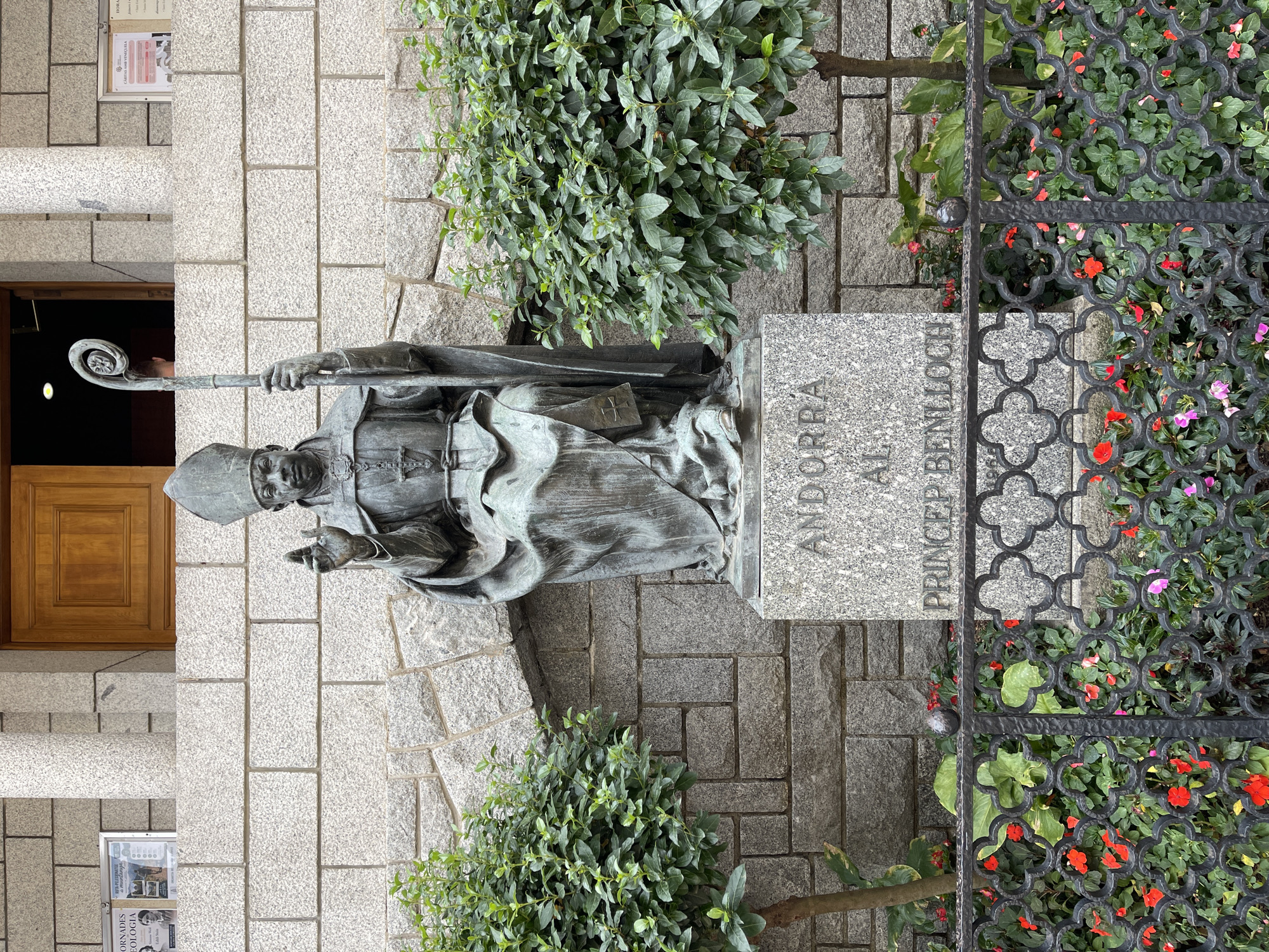 A full-shot, eye-level, medium outdoor photo shows a bronze statue of a seated bishop holding a crozier in his left hand and raising his right hand in a blessing gesture. The bishop wears a miter and vestments, and the statue sits atop a grey granite pedestal with the inscription "ANDORRA AL PRINCEP BENLLOCH". Two well-trimmed green bushes flank the statue, and a black wrought-iron fence with clover-like designs runs in front of them. Behind the statue, a stone building with a wooden door is visible. The scene is lit by overcast daylight.