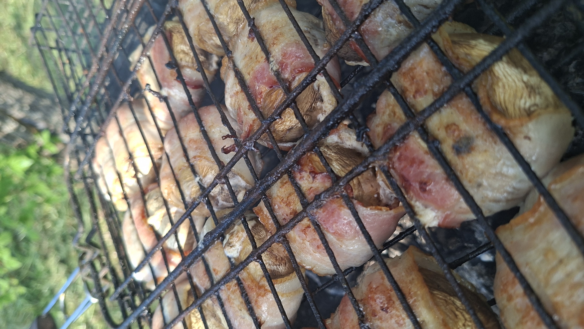 The image shows mushrooms wrapped in bacon grilling on a black metal grill. The grill appears to be outdoors, as evidenced by the green vegetation visible in the background. The focus is on the food, with the grill and background slightly blurred. There's some smoke present. The mushrooms are tan and appear mostly cooked, with some white parts still visible. The bacon is pink and appears to be sizzling. The image seems to be taken from a slightly elevated angle. The location is likely Сваткаўскі сельскі Савет, Belarus.