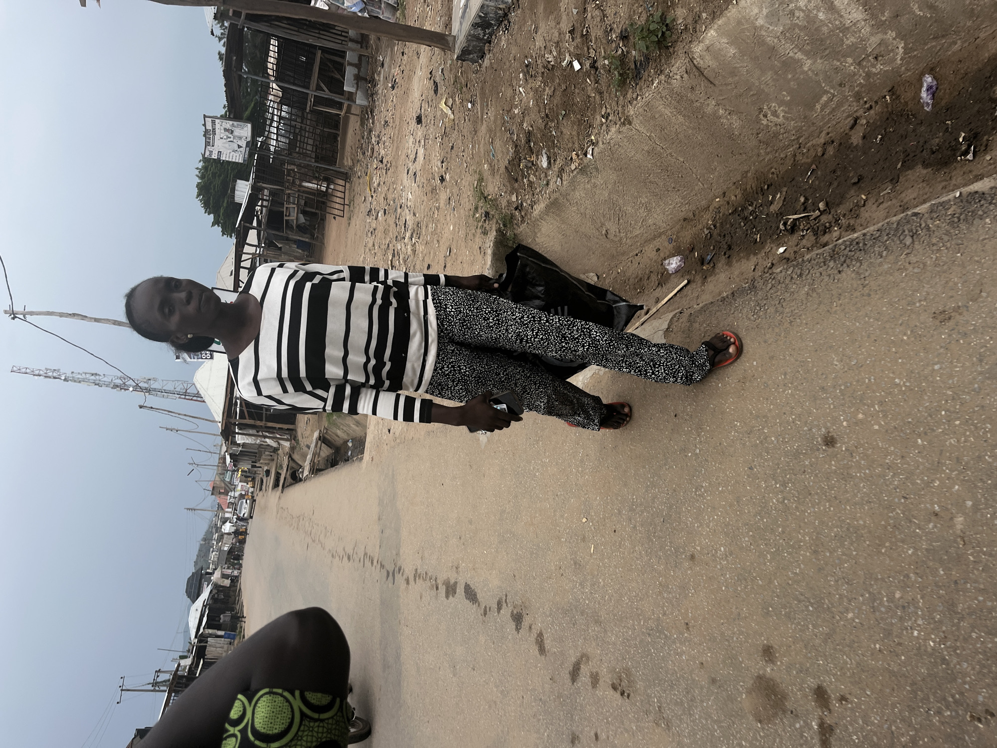 In Abuja, Nigeria, a woman walks down a dirt road lined with market stalls and a drainage ditch. She wears a striped long-sleeved shirt and patterned pants, carrying a black bag. The road is dusty, with some water stains and debris. In the background, buildings and a cell tower are visible under a cloudy sky.