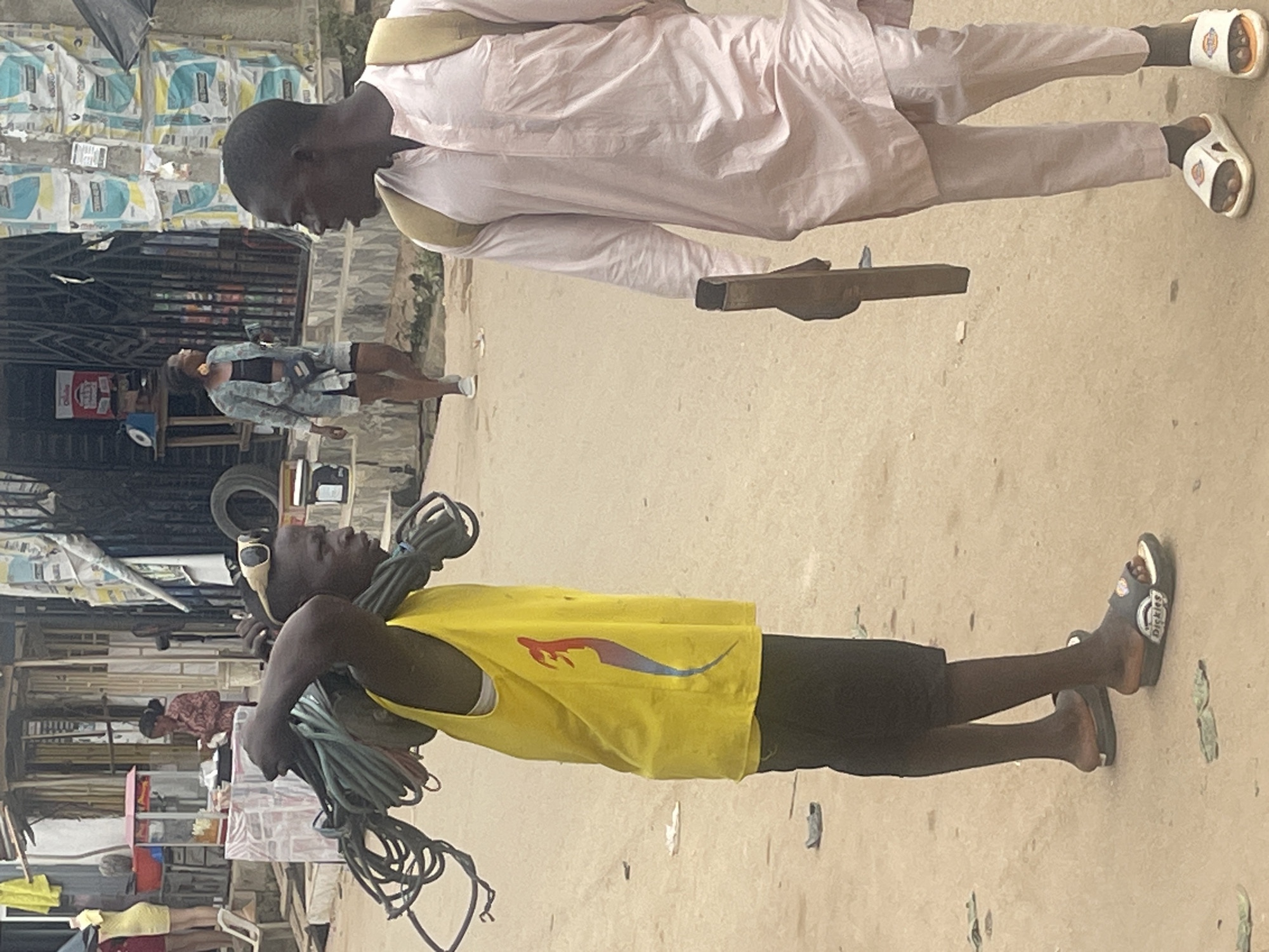 The image shows two individuals in an outdoor market setting. In the foreground, a person wearing a yellow tank top and black shorts is carrying several coiled bundles of what appear to be electrical cables or ropes. They are wearing sandals. In the background, another person is standing, wearing a light-colored, loose-fitting garment and white sandals. They are holding a rectangular wooden object.
The scene appears to be a busy street or market in Abuja, Nigeria. The ground is unpaved, with sand and dirt visible. In the background, there are stalls and structures that suggest a commercial area, with some goods displayed, including what looks like bags of cement or similar materials stacked against a wall. The lighting suggests it might be daytime, possibly morning or afternoon, with natural light illuminating the scene. There are no clear indicators of the weather, but the setting is consistent with a warm climate. The people's expressions are neutral, and the overall impression is one of everyday commerce and labor. Visible text is limited, but the sandals worn by the person in the foreground have "Dickies" written on them.