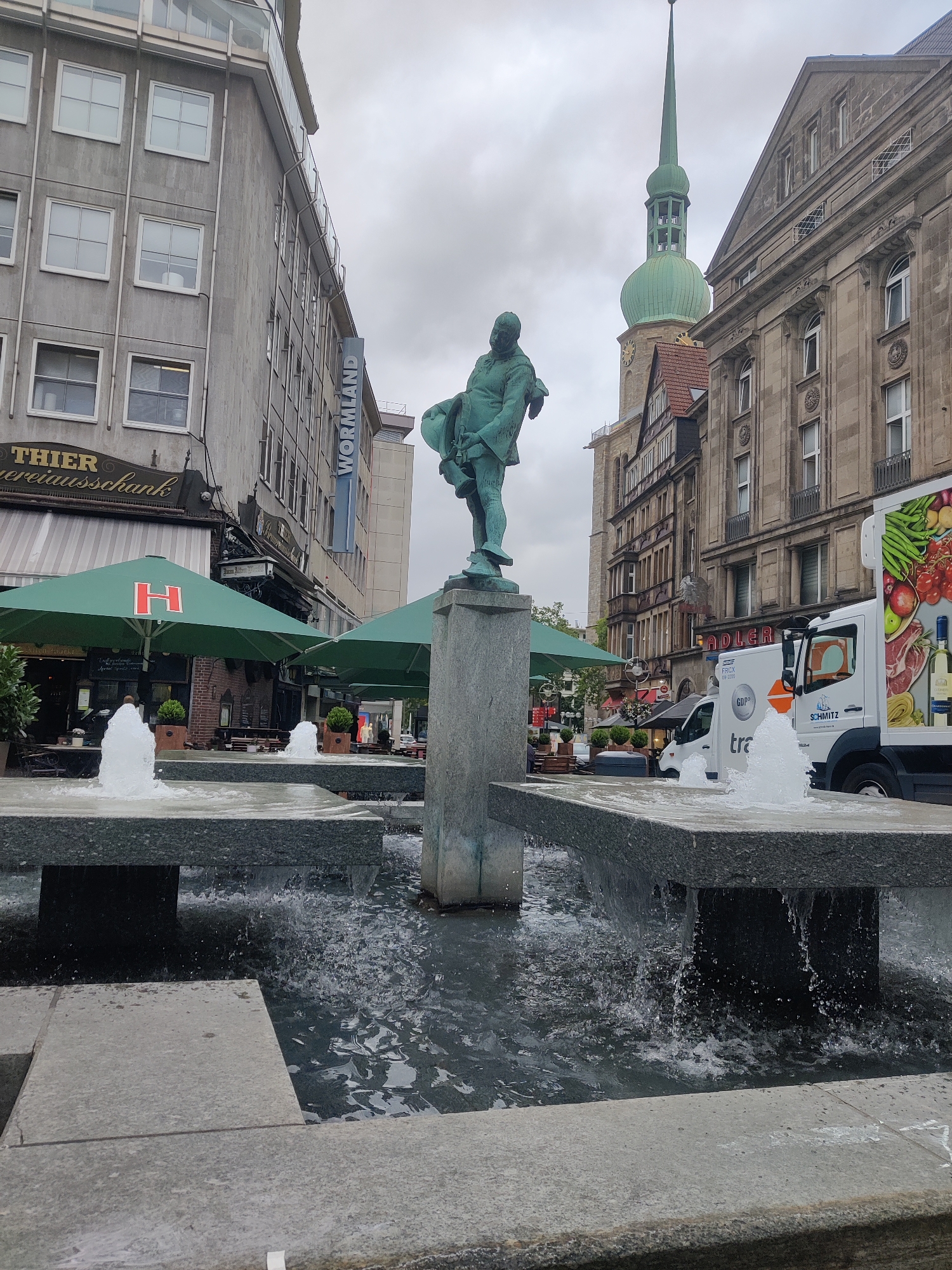 The image shows a street scene in Dortmund, Germany, specifically in the vicinity of the Thier Carousel or Thier Carousel Building. In the foreground, a multi-tiered fountain with granite-like surfaces is actively spraying water. A bronze statue of a man holding a basket is prominently featured on a pedestal in the center of the fountain area.
The background reveals an urban environment with several multi-story buildings. To the left, a gray building with numerous windows is visible, bearing the signage "THIER" and "vereinsbank". Adjacent to it, a blue vertical sign reads "WÖRMLAND". Further in the background, a green-spired church tower is visible. To the right, another building with stone facade and arched windows stands. A white delivery truck with an advertisement for fresh produce and other goods is parked on the street. The sky is overcast. Several green outdoor umbrellas are set up, suggesting a cafe or restaurant seating area. No individuals are clearly discernible or engaged in specific actions within the frame. The overall impression is of a public square or pedestrian area in a European city.