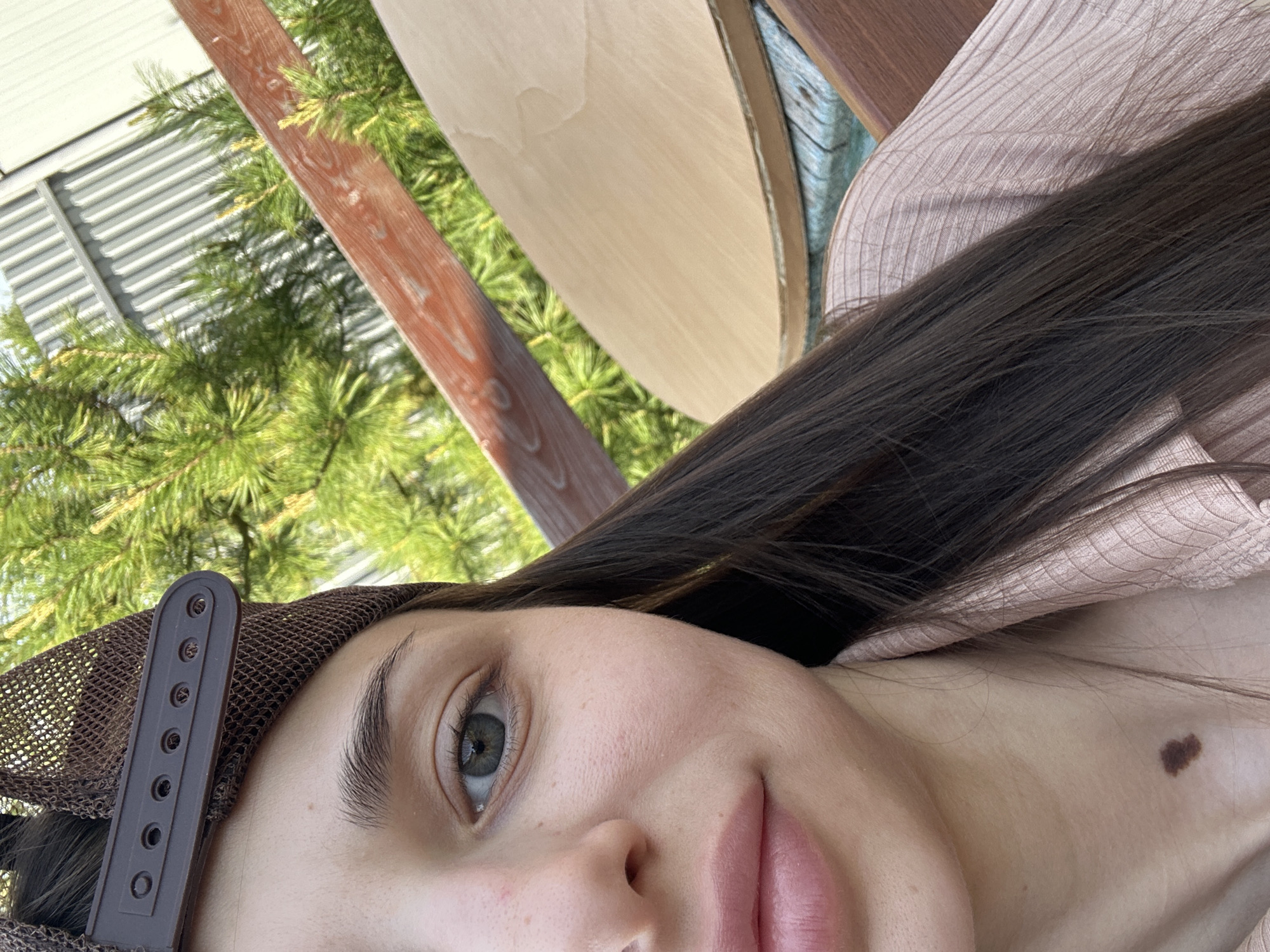 A close-up, tilted perspective photograph shows a young woman's face and upper torso. She has light skin, dark brown hair, and light-colored eyes. She wears a brown mesh baseball cap. A large mole is visible on her upper chest. The background includes green foliage, what appears to be a metal corrugated structure, a wooden beam, and a light-colored wooden surface. The location, Ludorvai, Russia, cannot be verified by the visual information alone.