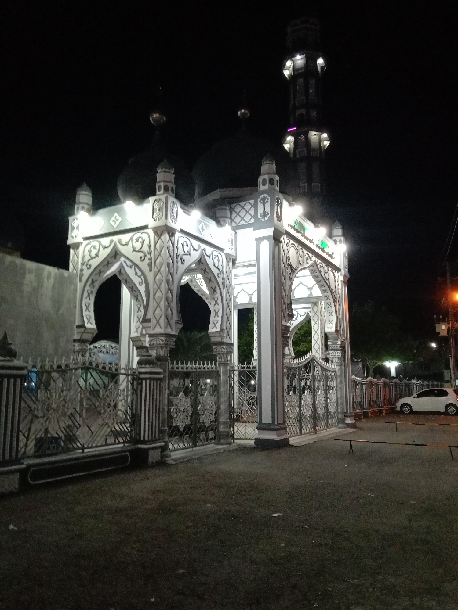 Captured at night in Keudekruenggeukueh, Indonesia, the image depicts a mosque entrance illuminated against the dark sky. The structure consists of a white and gray archway adorned with intricate patterns and flanked by decorative gates. The mosque's domes and minaret are visible in the background, also lit up. A white car is parked on the right side of the image, near a barrier. The foreground shows a paved area.
