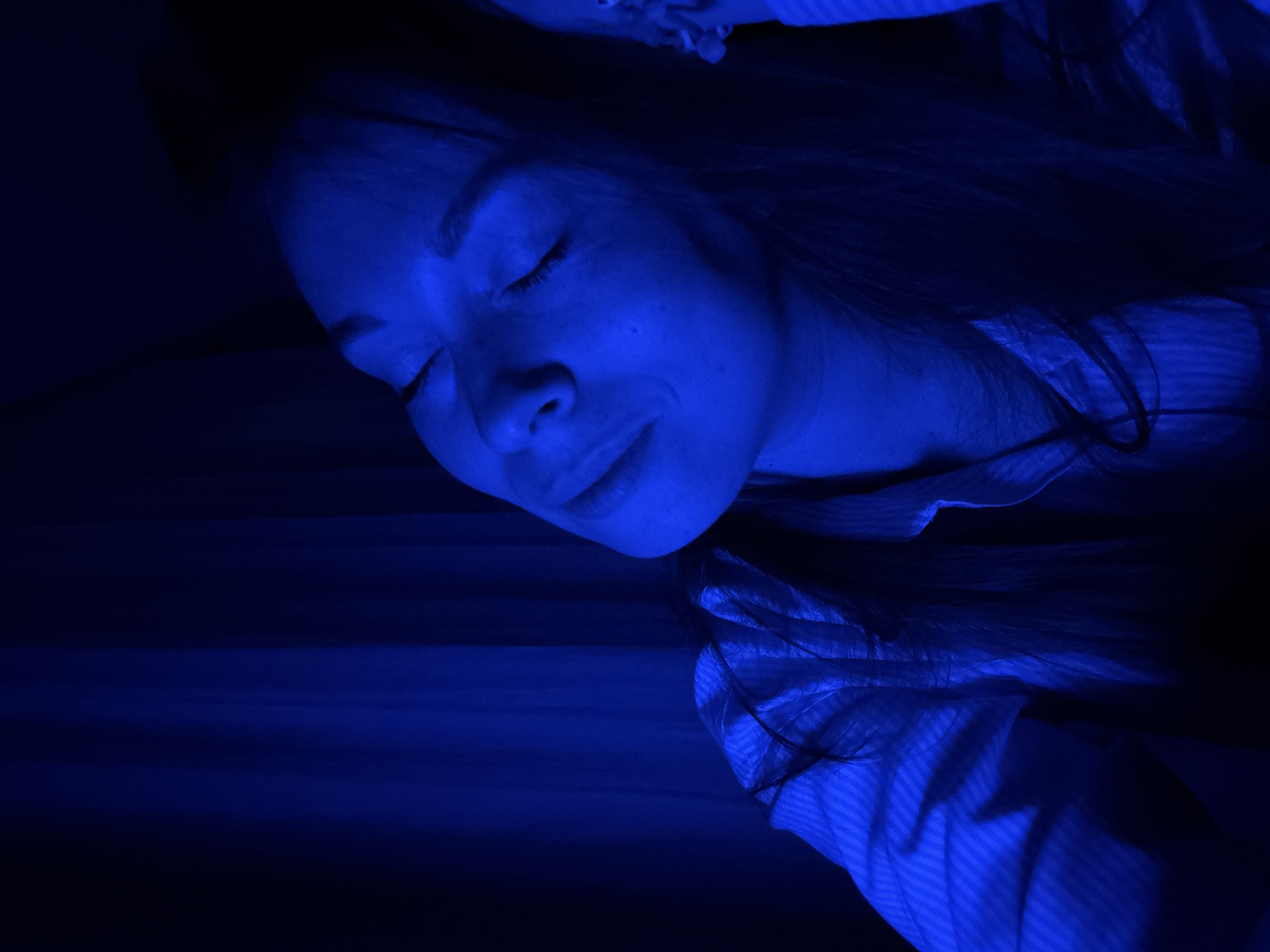 The image shows a woman indoors, likely in Budva, Montenegro given the prompt's context. She is lying down with her eyes closed, possibly asleep. The scene is illuminated by a strong blue light. She has long dark hair and is wearing a striped shirt. The background is dark and difficult to discern specific details.