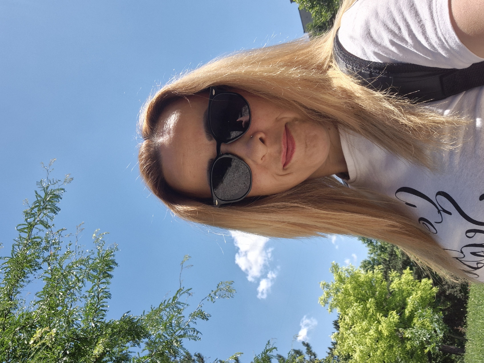 A woman with blonde hair and sunglasses takes a selfie outdoors on a sunny day. She is wearing a white t-shirt with black lettering. The background shows a clear blue sky with some clouds, green trees, and a hint of a building.