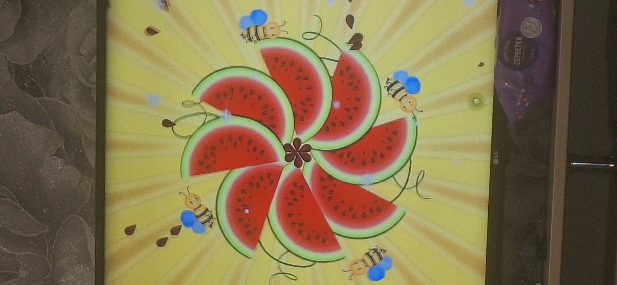 A digital screen displays a cartoon image of a sunflower made of watermelon slices, with bees flying around it. The screen is placed on a shelf in front of a textured gray wall. In the foreground, a purple package with Russian text that translates to "SERBETSKI" is visible. The overall scene suggests a domestic setting, possibly a living room or entertainment area. There is no discernible information to locate this image within the city of Bila Tserkva, Ukraine.