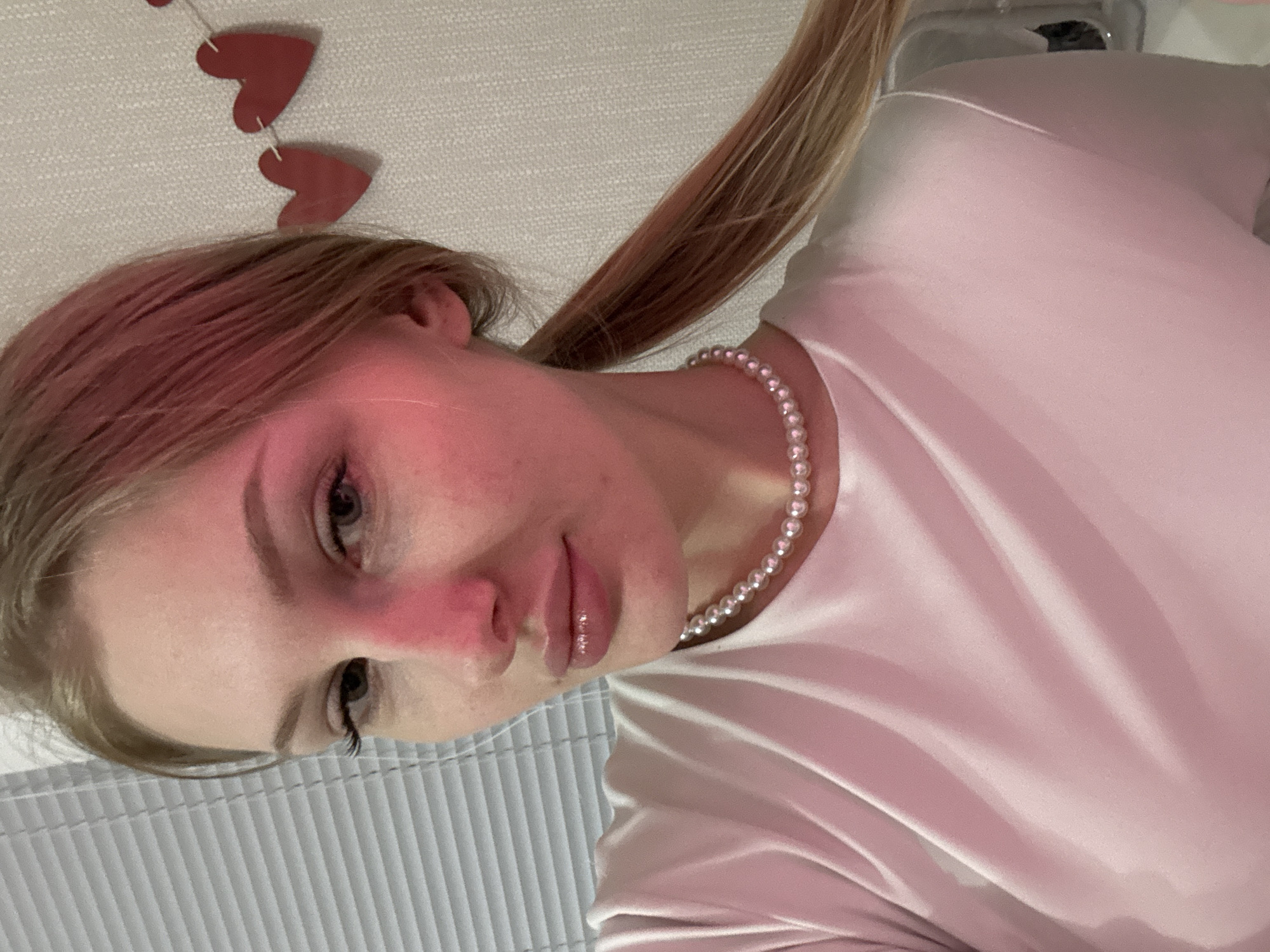 The image is an indoor portrait of a young woman, likely a selfie, taken in Perm, Russia. She has light-colored, possibly blonde or light brown hair, which is pulled back into a ponytail visible on the right side. Her complexion is fair, with a noticeable flush on her nose and cheeks. She has light-colored eyes and full lips with a glossy finish.
The woman is wearing a light pink, possibly satin or silk-like, short-sleeved top. Around her neck, she has a white pearl necklace. Her head is tilted, and she is looking slightly downwards towards the camera with a calm, neutral expression.
The background features a light-colored, textured wall. Towards the upper left, two red heart-shaped decorations are hanging, suggesting a decorative or festive theme, possibly related to Valentine's Day. In the lower part of the image, below her head, a gray, ribbed surface can be seen, which appears to be either a radiator or window blinds. The lighting is soft and warm, indicative of indoor artificial illumination. There is no visible text in the image. The overall scene conveys a personal and relaxed moment indoors.