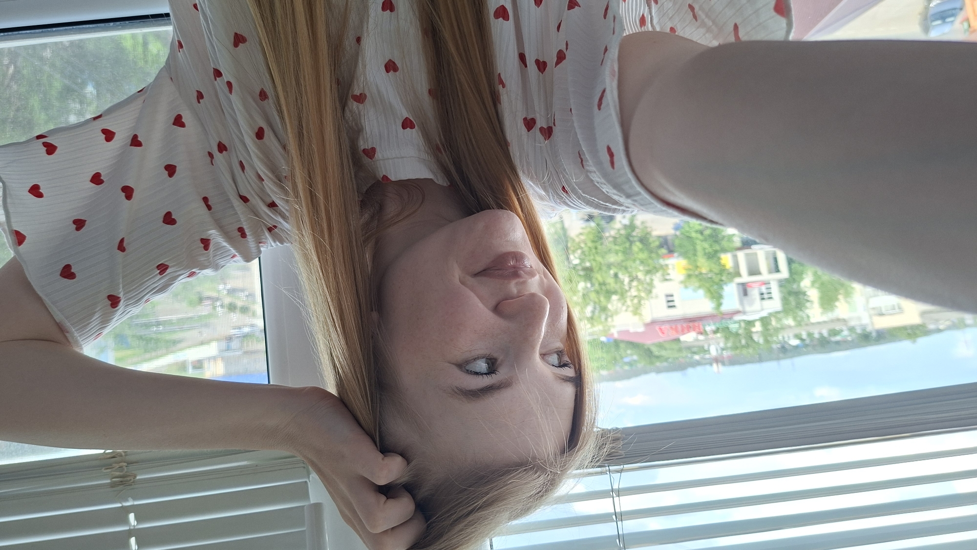 A fair-skinned person with red hair is upside down in front of a window with white horizontal blinds. The person is wearing a white shirt with red heart-shaped prints. The background shows a cityscape, with trees and buildings visible through the window. Based on the visible buildings and landscape, the location is Chaikovsky, Russia.