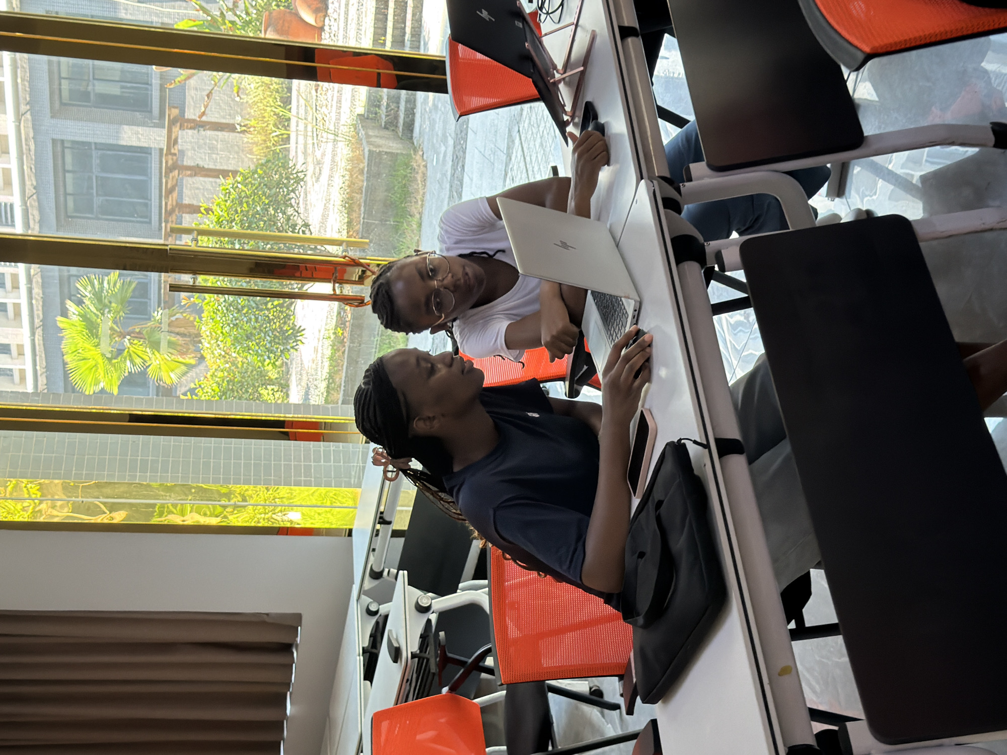 Two young Black women, likely students, are seated at white desks in a bright, modern classroom or study area with large windows. One woman, on the left, wears a dark blue t-shirt and has her hair in braids. She is looking at a laptop in front of her, with her right hand on a mouse. On the desk in front of her is a black laptop bag and a pink smartphone. The other woman, on the right, wears a white t-shirt and glasses. She is leaning forward, looking intently at the screen of her laptop, which is open on a rose-gold colored laptop stand. Her left hand is on her chin, and her right hand is resting on the desk. Another laptop is open on a stand to her right. The background shows a contemporary outdoor space with greenery, palm trees, and buildings visible through the windows. The desks have a white top and white metallic legs, and the chairs are bright orange with black mesh backs. The flooring appears to be polished concrete or tile. The scene suggests an educational or collaborative work environment. The location is identified as Jiangjin, China.