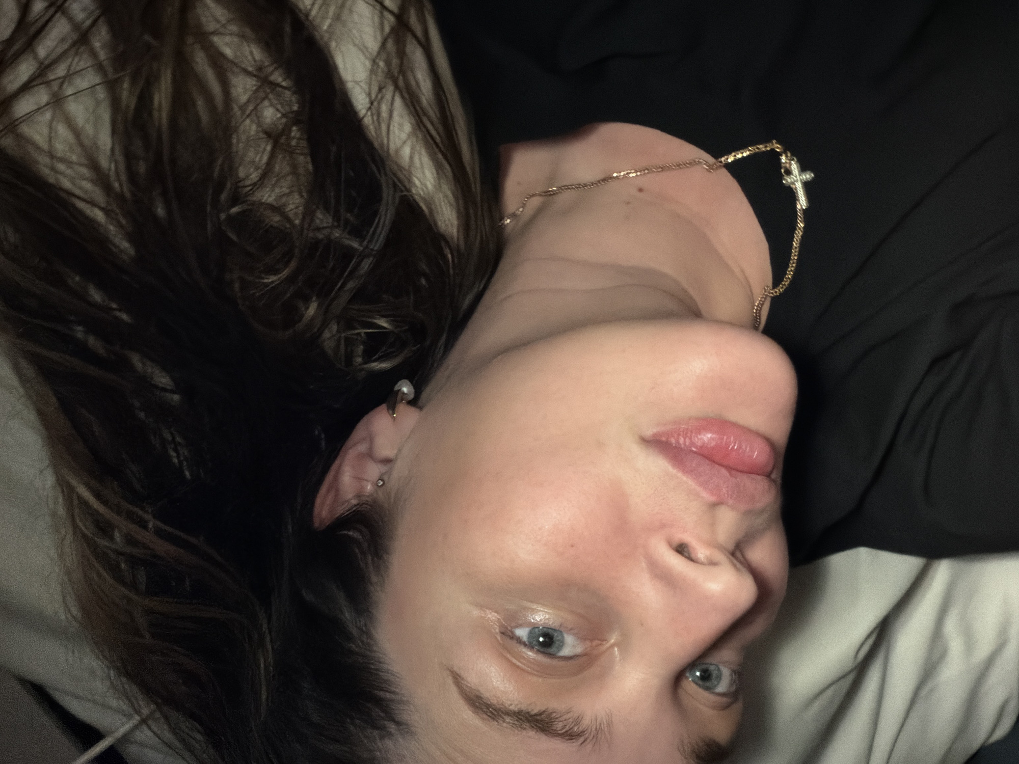 This media file is a close-up portrait of a young woman, rotated ninety degrees counter-clockwise from a standard upright view. The woman has fair skin, light blue eyes, and full, slightly parted lips, expressing a neutral or calm demeanor as she looks directly at the camera. Her long, dark brown hair, with visible lighter streaks, is fanned out around her head, adding texture to the left side of the frame.
She is wearing a thin, gold-colored chain necklace with a small, silver-colored cross pendant resting on her neck. Her left ear is visible, adorned with a small stud or pearl-like earring in the lobe and a gold-colored hoop or cuff piercing in the upper cartilage.
The background suggests an indoor setting, likely a bedroom. A crumpled white or light-colored sheet or pillow is visible on her left side (bottom right of the rotated image), while a dark, possibly black, fabric is seen on her right (top right of the rotated image). The lighting is soft and appears to be artificial or diffuse. The image captures a personal moment, possibly a casual selfie, emphasizing her face and upper body. No text is visible within the image. The provided location context is Moscow, Russia.