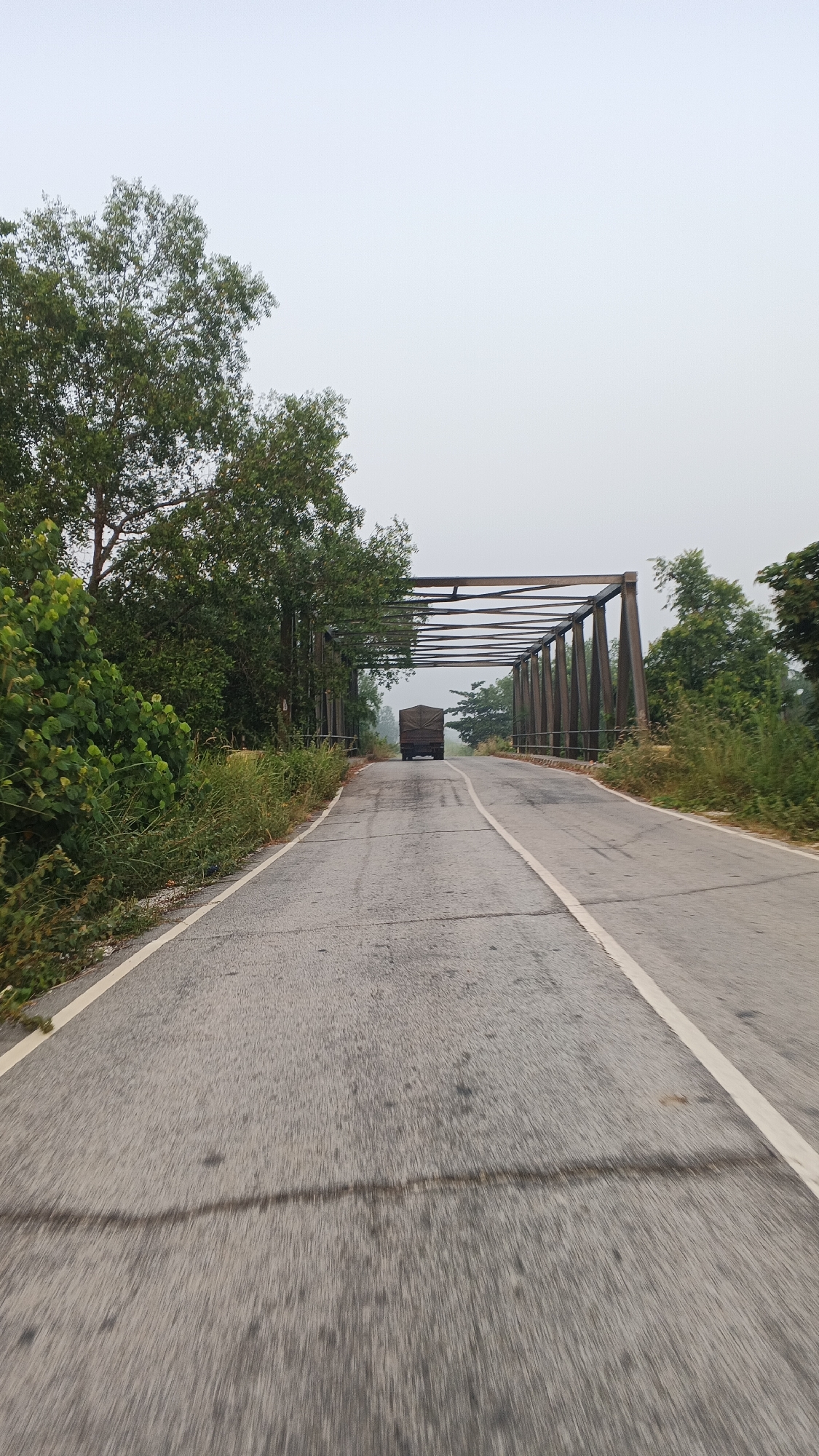 A truck is driving on a road that leads to a bridge. The road is asphalt and has white lines. The bridge is made of metal and has a truss structure. There are trees and vegetation on both sides of the road. The sky is overcast. The scene is located in Rawang Air Putih, Indonesia.