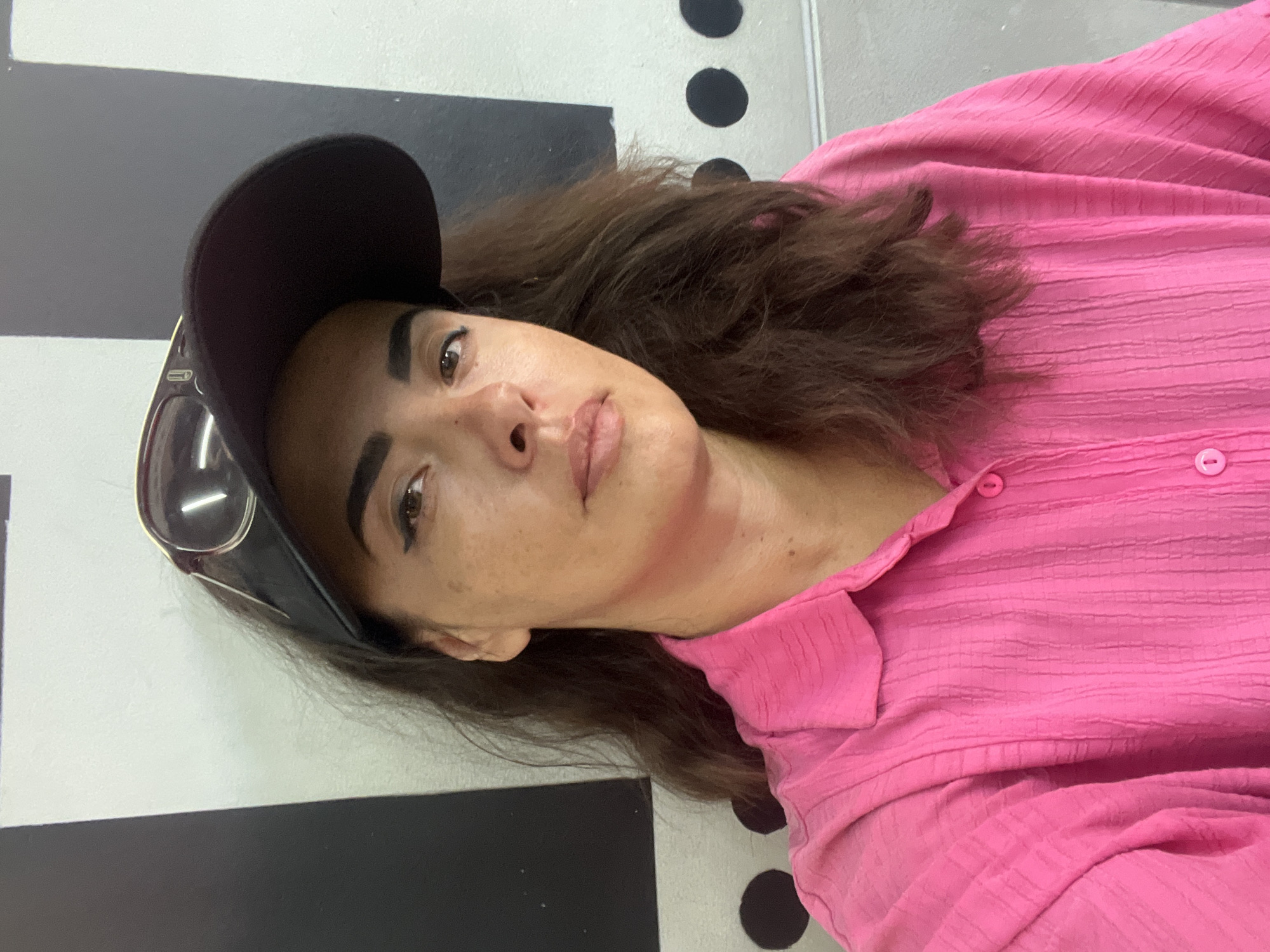 A medium shot captures a woman wearing a bright pink, textured button-down shirt and a black visor with sunglasses resting on top. She is looking directly at the camera with a neutral expression. Her dark hair is loose and falls over her shoulders. The background consists of a wall with a black and white geometric pattern and some black circles. The image appears to be taken indoors, possibly in an urban setting like Kuta Selatan, Indonesia, based on common architectural styles.