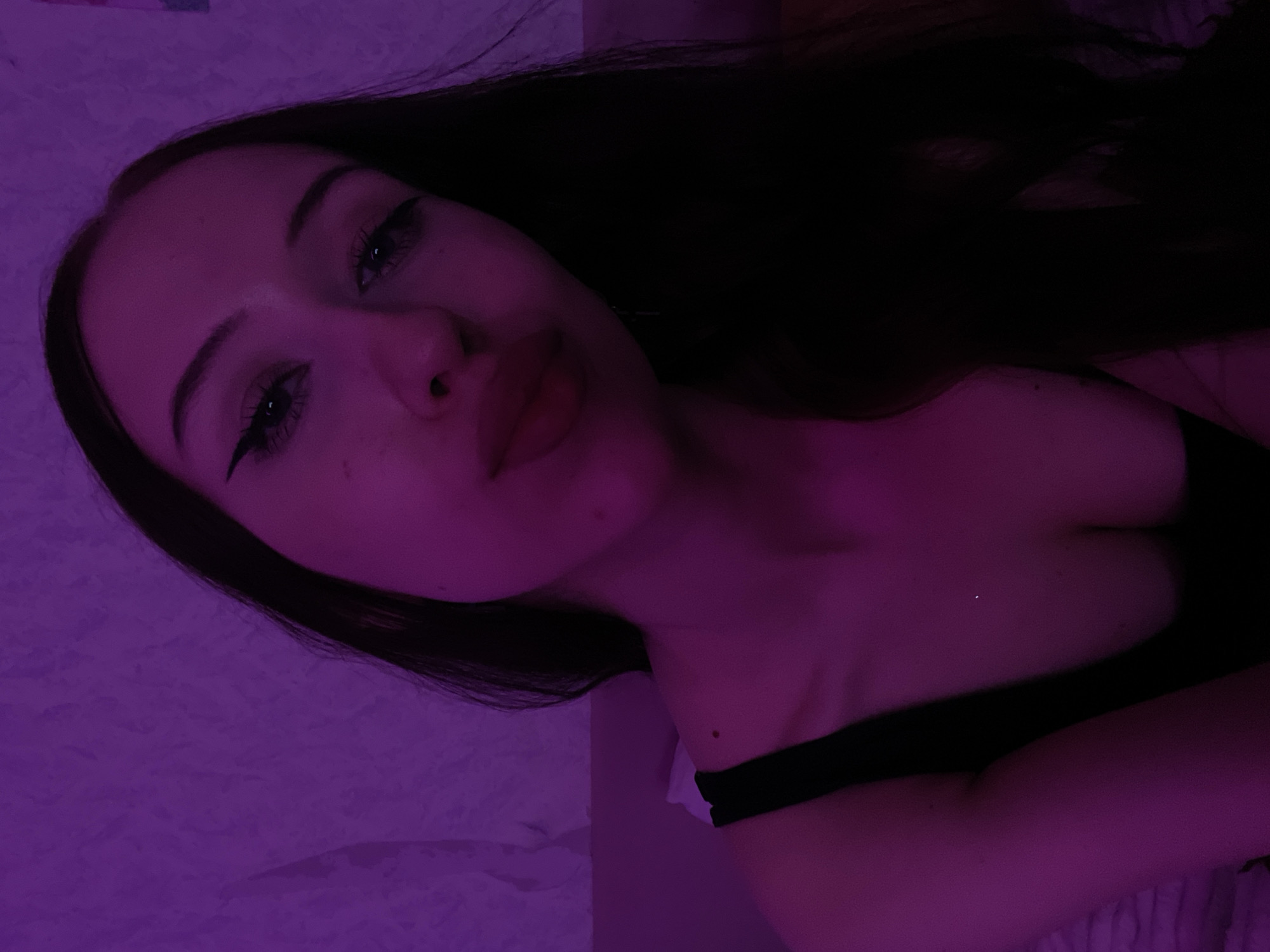The image shows a close-up, chest-up selfie of a young woman with long, dark hair. She is wearing a black top and has on eyeliner. The lighting is predominantly purple, casting a violet hue over the scene and creating a textured background that appears to be a wall or fabric. The woman is looking towards the camera with her lips slightly parted. No specific location in Votkinsk, Russia is identifiable from the image.