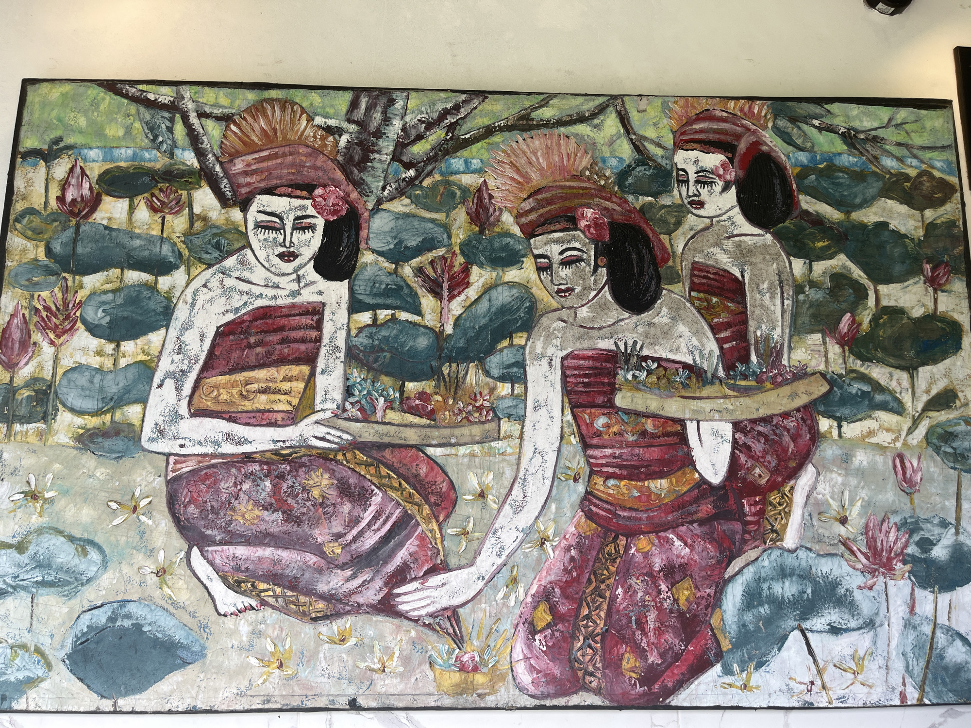The image is a painting depicting three women in traditional Balinese attire, kneeling amidst a lotus pond. The women wear red and gold clothing, head coverings with feathered accents, and heavy makeup. Each woman carries a tray containing offerings of fruit and flowers. The backdrop consists of lotus leaves, lotus flowers, and a tree branch against a muted green and blue sky. The style of the painting is primitive and textured. The location is likely near Tegallalang, Indonesia, given the traditional Balinese attire.