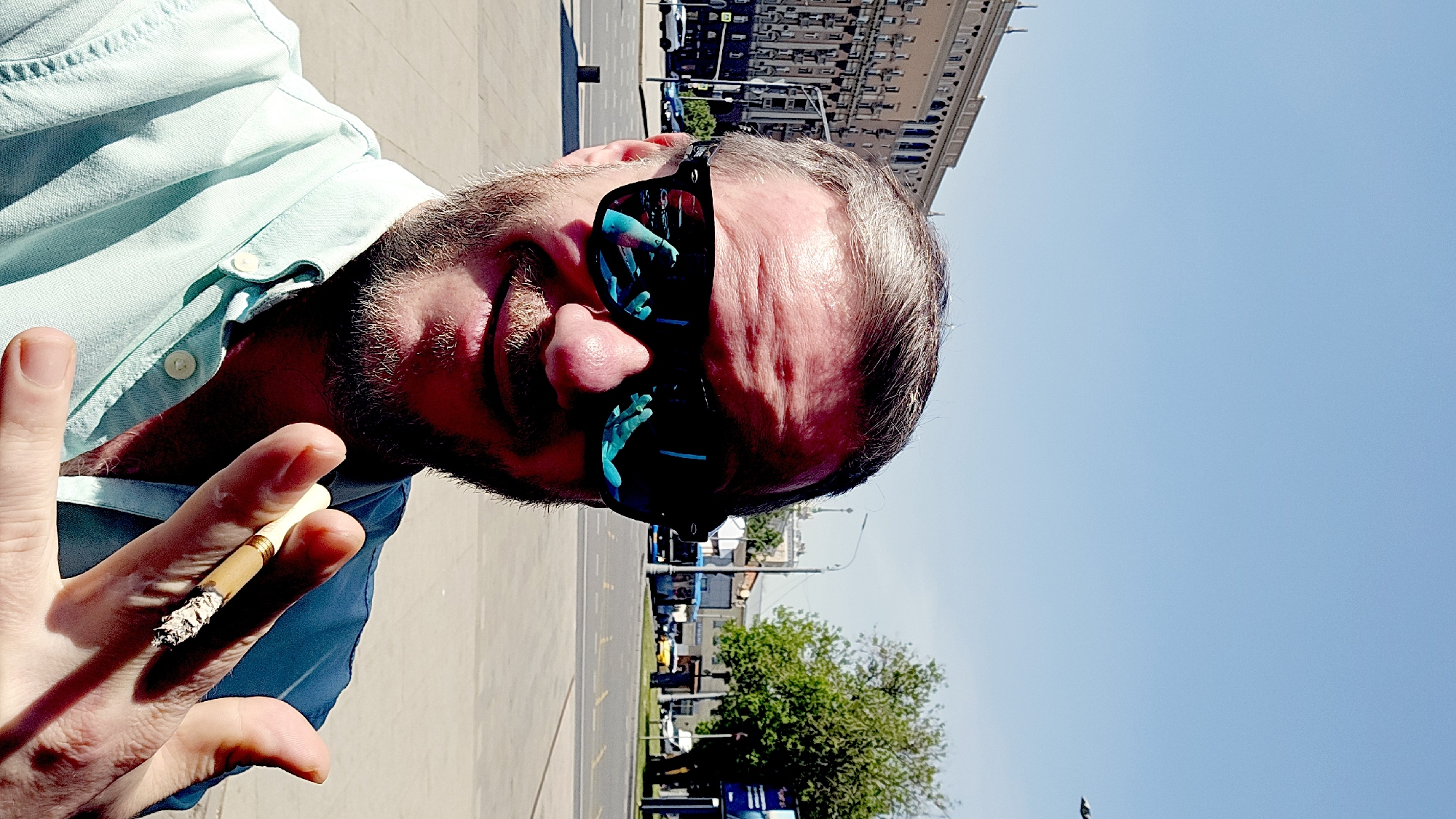 A man with light skin and a beard is seen in a selfie outdoors. He wears sunglasses with blue reflections, and a light blue button-down shirt. He is holding a lit cigarette between his fingers. The background shows a city street with trees, cars, and buildings, suggesting an urban setting. Based on the architecture and layout of the scene, the location is plausibly in Moscow, Russia.