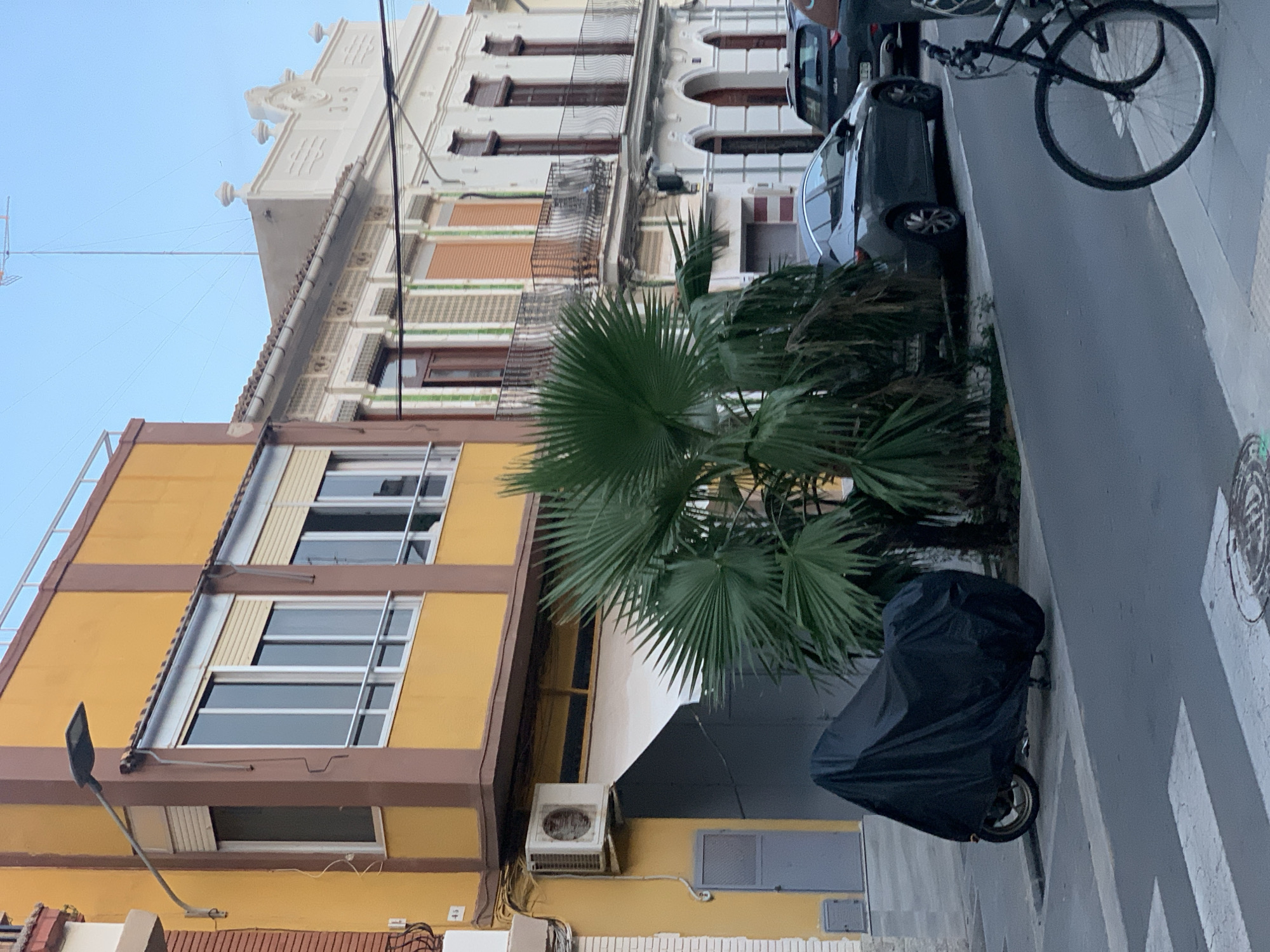 This is a medium shot, eye-level, street-level photograph taken from the perspective of the sidewalk. The scene depicts a residential street in the city of Valencia, Spain, with a clear blue sky and a few wisps of clouds.
In the foreground, a pedestrian crossing is visible. To the left, a motorcycle covered in a black tarp is parked parallel to the curb. Next to the motorcycle is a dark green palm tree with large, fanned leaves. Behind the palm tree, several cars are parked on the street, including a dark gray sedan and a darker SUV. A bicycle with a black frame and silver wheels is also parked on the sidewalk to the right of the frame.
The background features a row of multi-story buildings. The building on the left is painted yellow with brown trim and has several windows with rolling shutters and a balcony. An air conditioning unit is mounted on the lower section of this building. To its right, a white building with ornate architectural details, including balconies and decorative elements, is visible. This building has windows with dark frames and wooden shutters. Further down the street, another white building with arched doorways and decorative molding is present. Electrical wires and a tall antenna are visible against the sky above the buildings. The overall impression is a residential street in a European city, likely during daylight hours, with a mix of older and more modern architectural styles.
