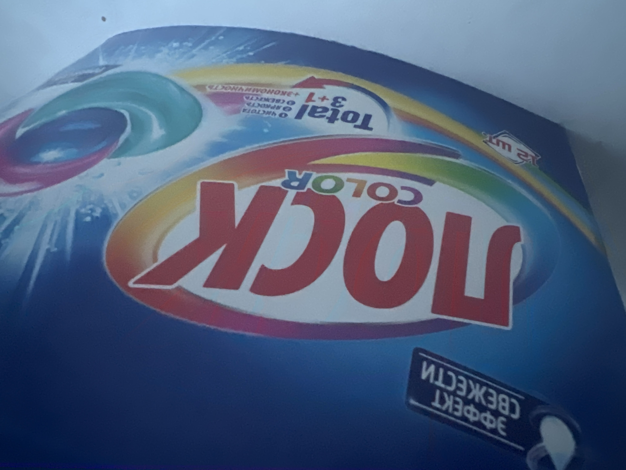 The image displays a close-up of a product package, likely for laundry detergent pods. The dominant text on the package is "ЛОСК COLOR" in large red and white lettering. Above this, a smaller black label reads "ЭФФЕКТ СВЕЖЕСТИ" (Effect of Freshness) with a graphic of a water droplet. Below "ЛОСК COLOR," there is a section with text that includes "Total 3+1" and other smaller text. In the lower left corner, a small red and white label shows "12 шт." (12 pieces). The background is a vibrant blue with stylized water splashes and rainbow-colored arcs. There are also colorful representations of laundry pods on the right side of the package. The image is shot from a slightly elevated angle, with the product taking up most of the frame. There is no discernible location in Moscow, Russia, or any people present. The scene is a still-life product shot.